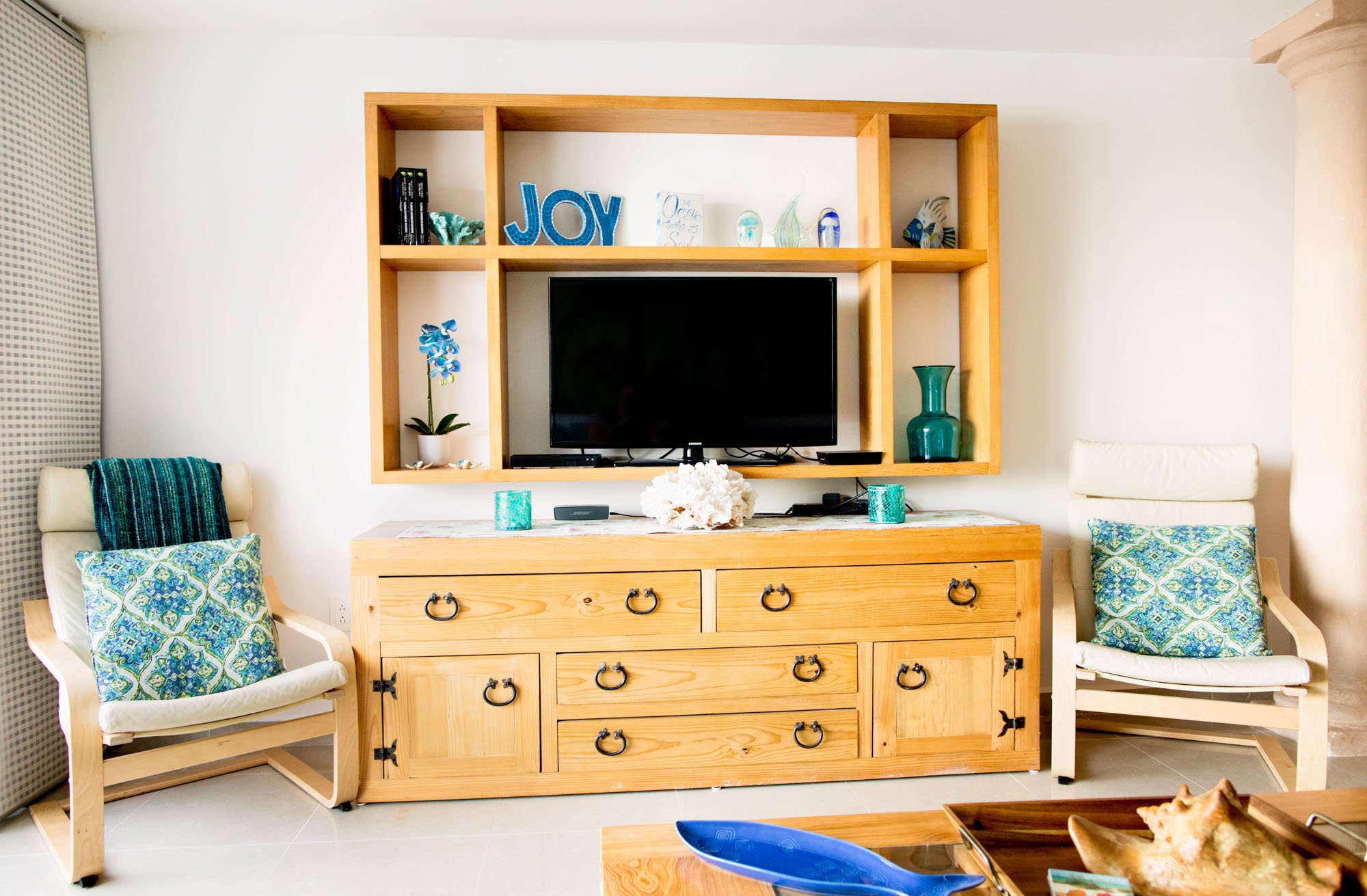 Living room with a wooden TV stand, flat-screen TV, and chairs with pillows, decorated with coastal-themed decor.