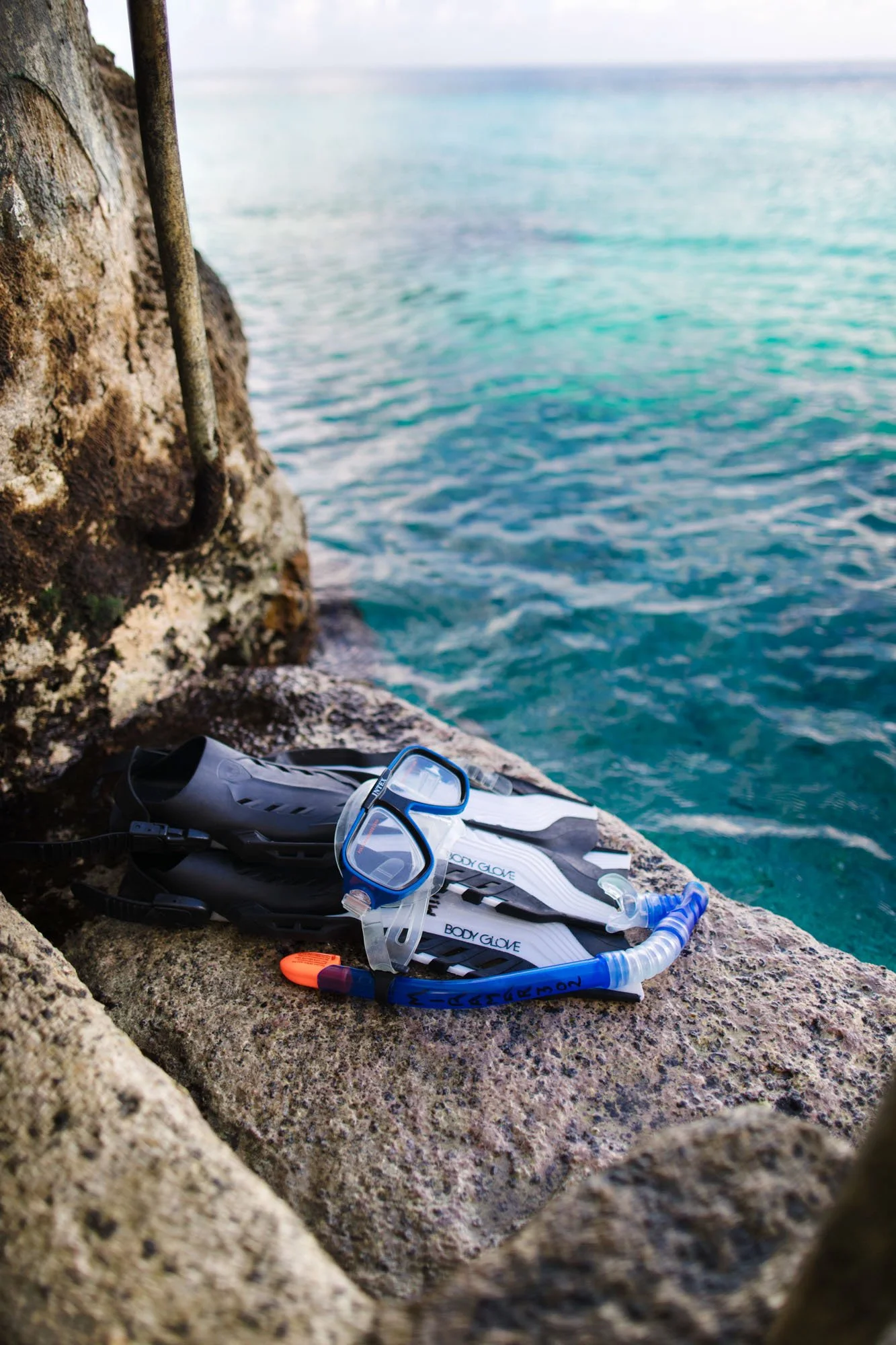 Snorkeling gear, including a mask, snorkel, gloves, and fins, resting on a concrete ledge by the water at a rocky shoreline.