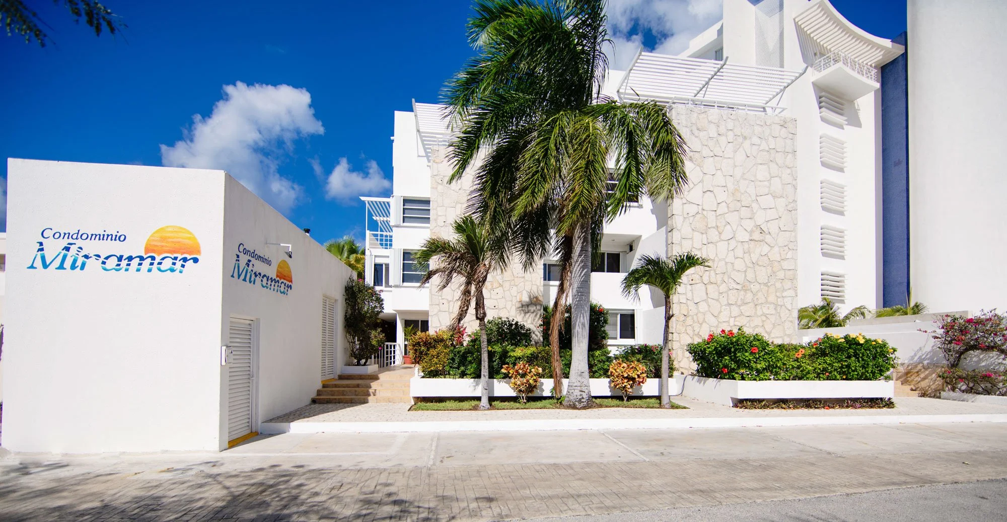 Exterior view of Condominium Miramar with white building, colorful flowers, and palm trees under a blue sky.