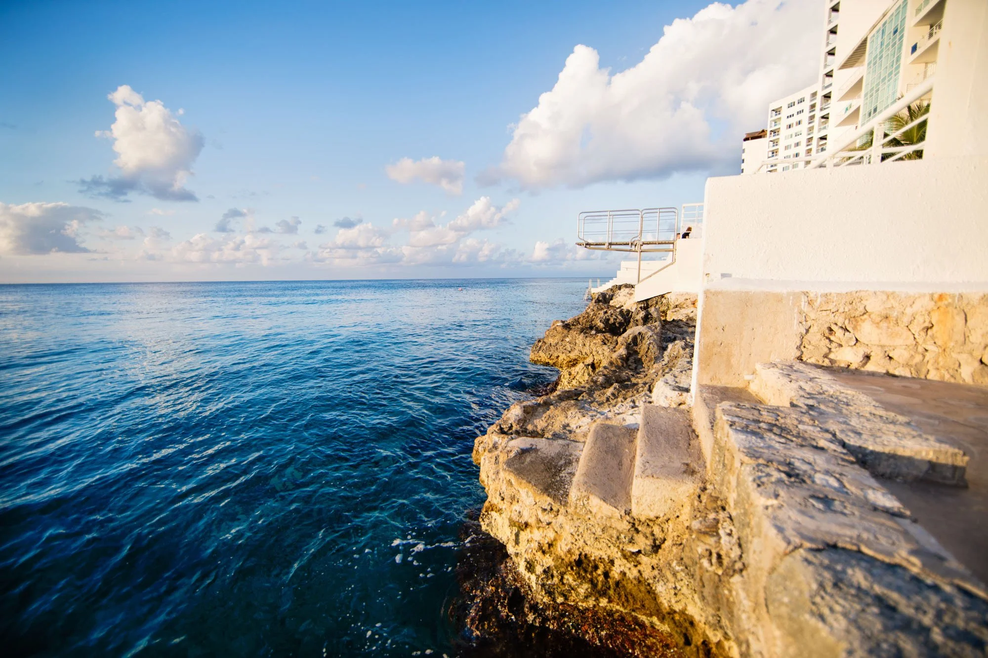 A view of the ocean with calm blue water, a rocky shoreline with steps, and a white building with balconies on the right, under a partly cloudy sky.