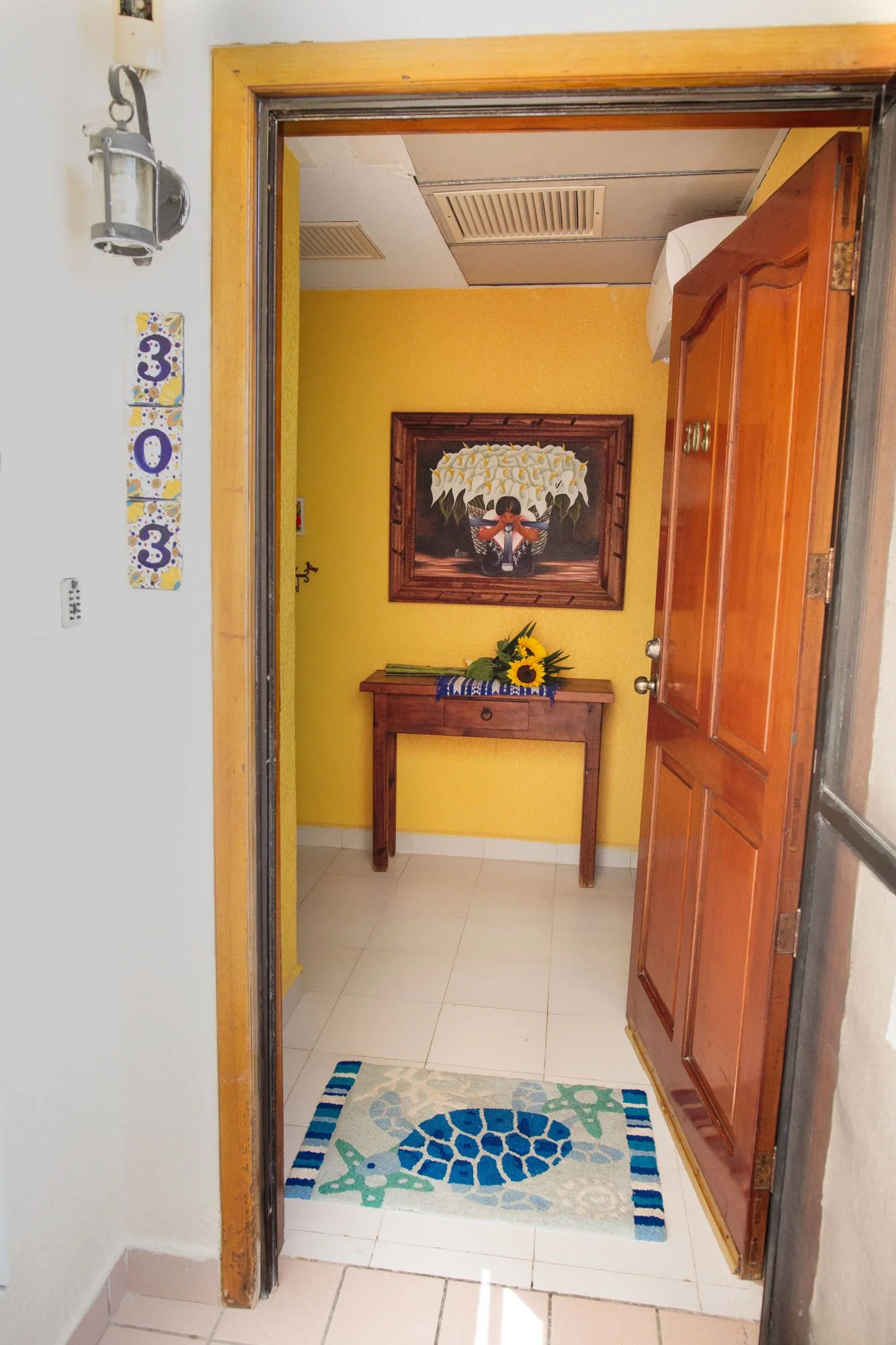 View of a small entryway with a yellow painted wall, a wooden console table with a sunflower flower arrangement,  and a framed floral painting above it. The open wooden door reveals a tiled floor with a turtle-themed rug in front.