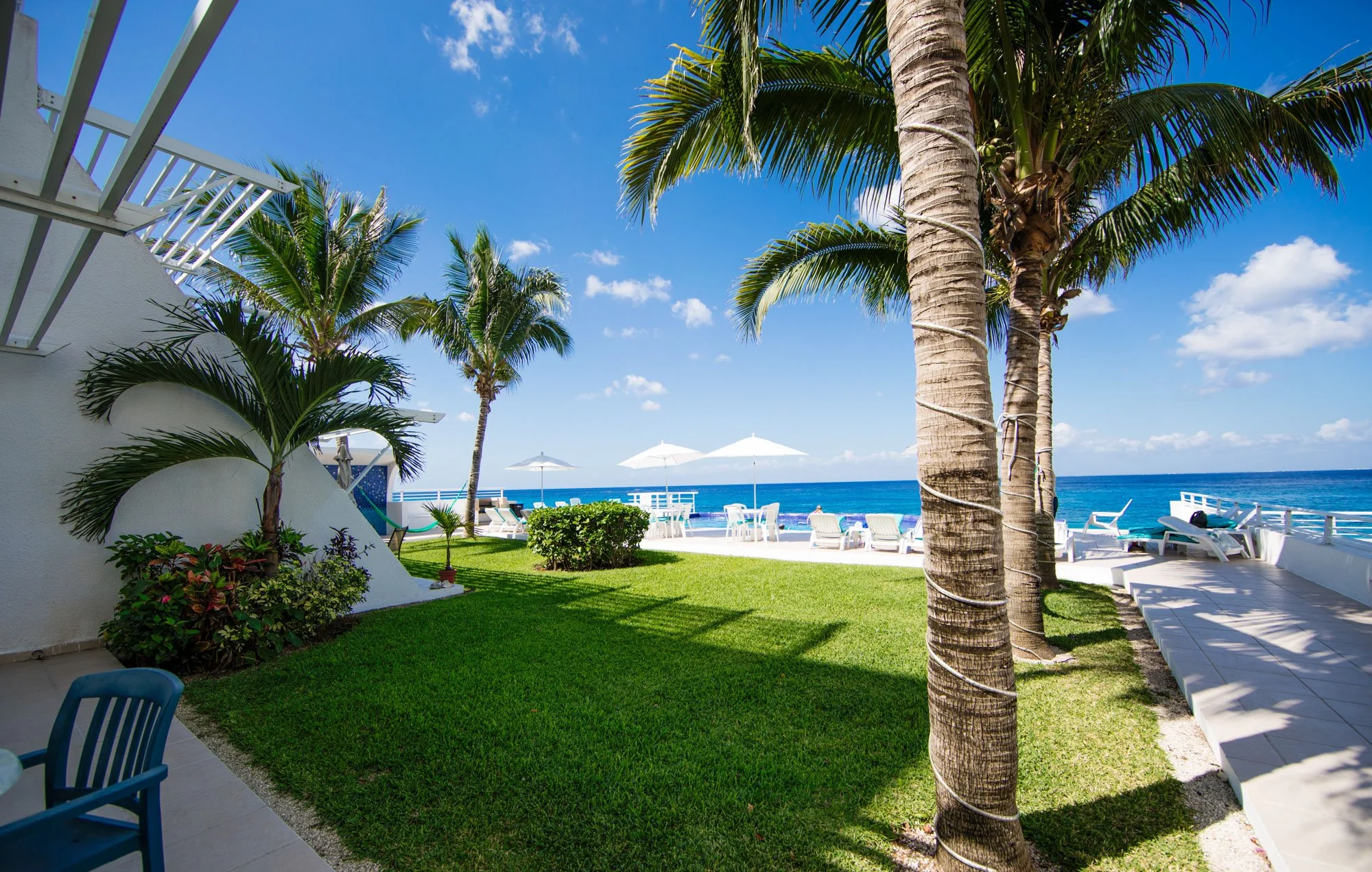 Beachside Cozumel vacation rental outdoor patio with palm trees, lawn, lounge chairs, tables with umbrellas, ocean view, clear sky.