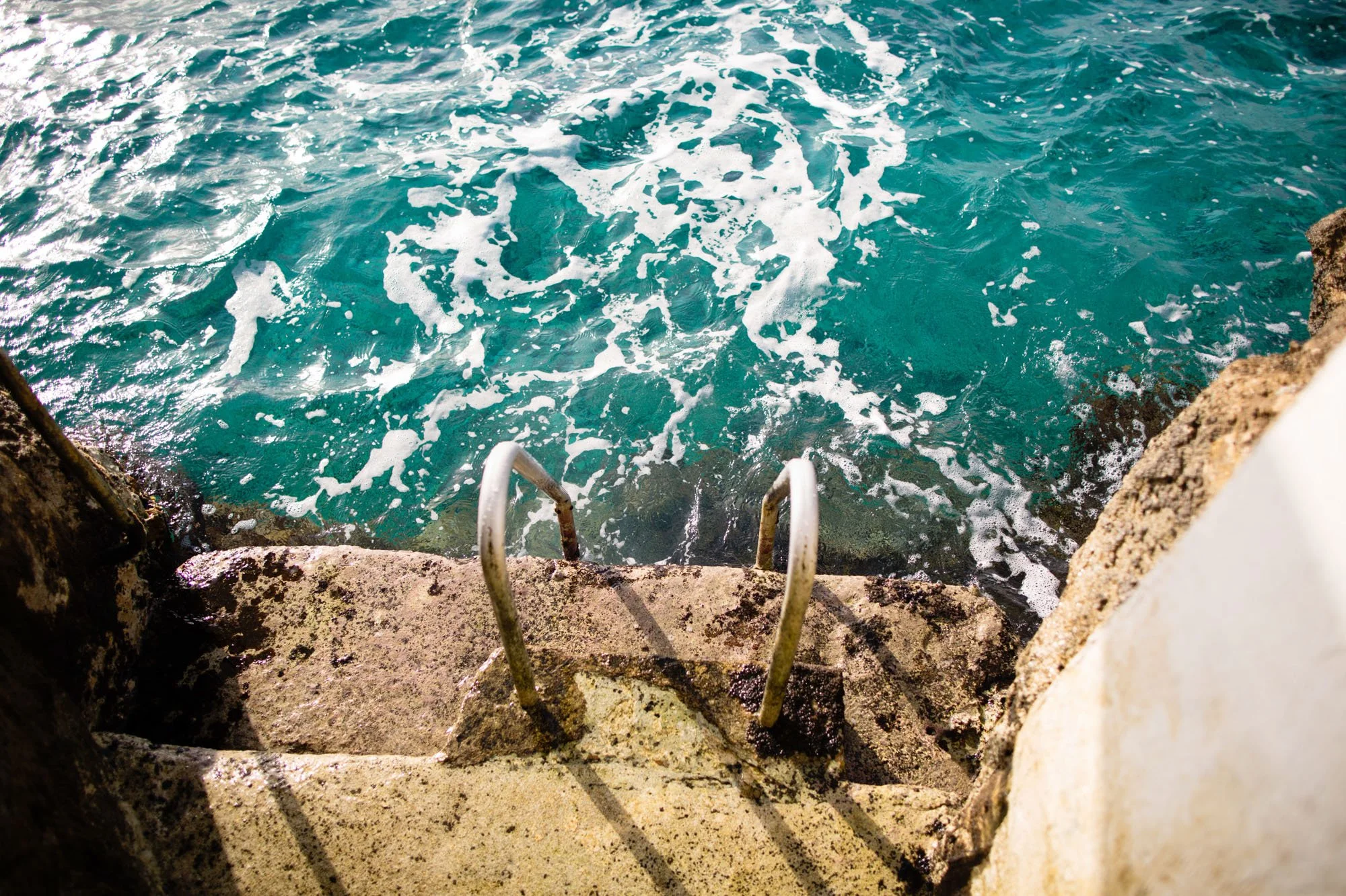 A view looking down at the ocean with turquoise waters and foam, seen from a concrete dock with metal handrails.