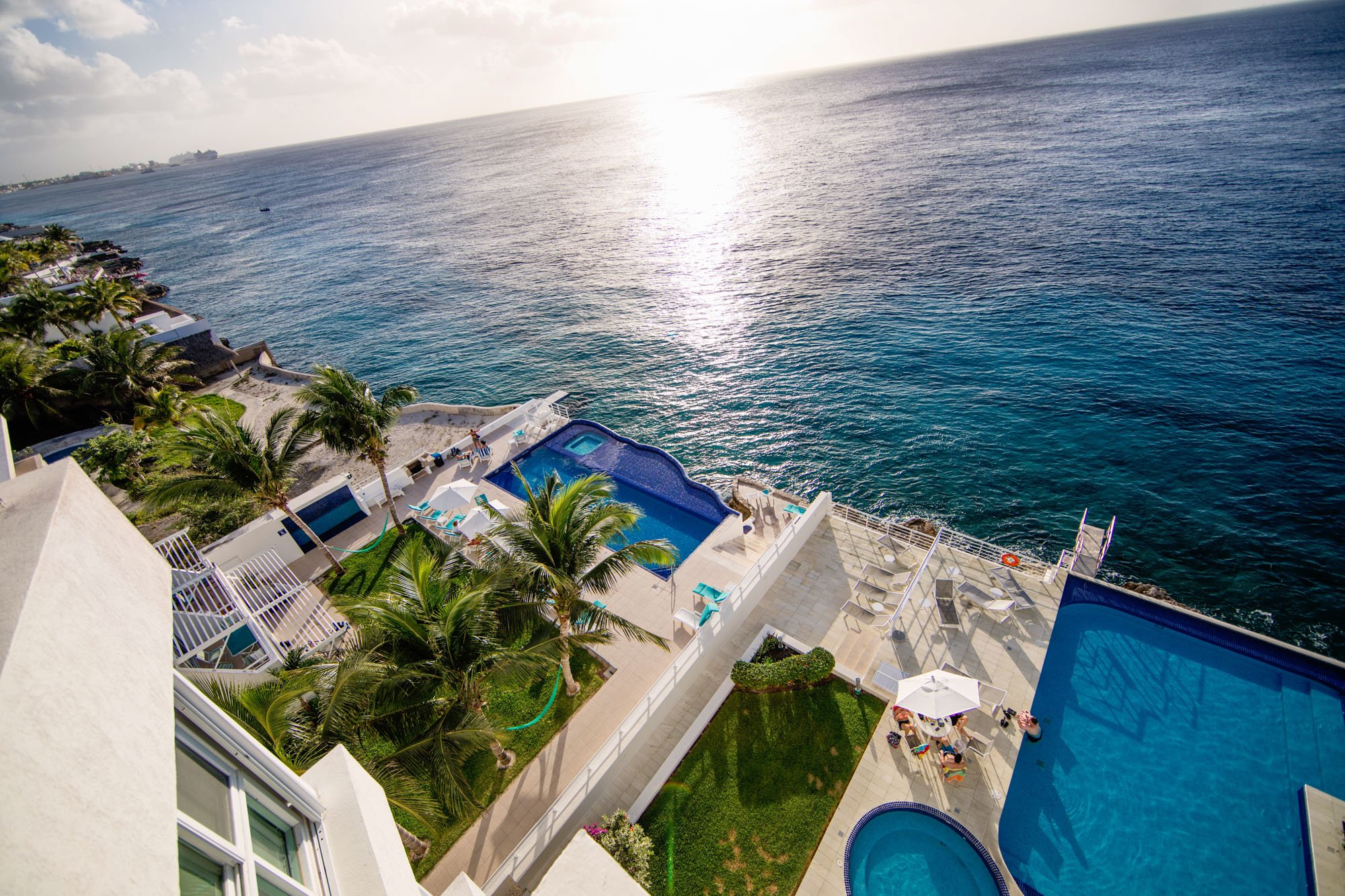 Aerial view of a Cozumel vacation rental with swimming pools, lounge chairs, palm trees, and a large ocean in the background during sunset.