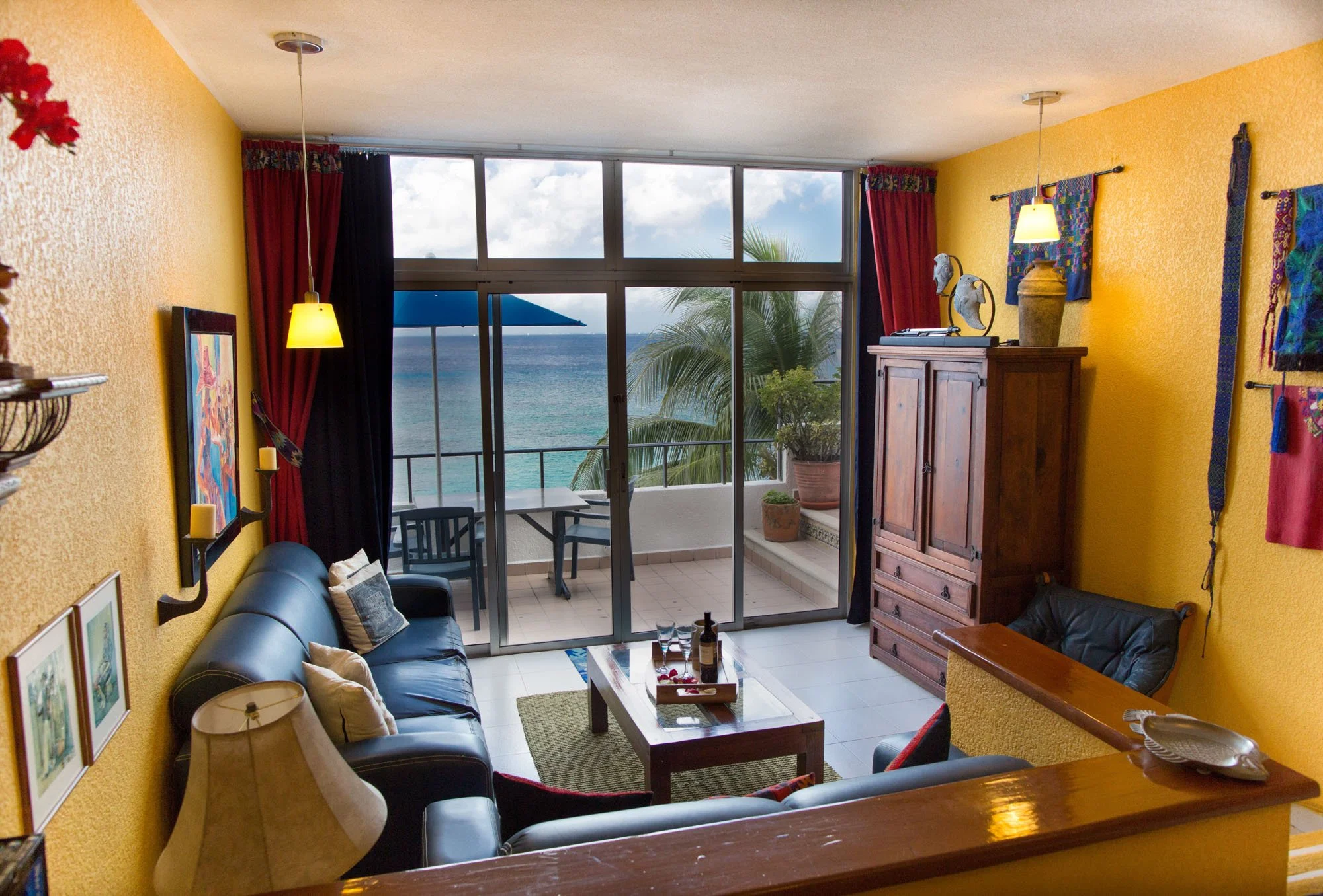 Cozumel vacation rental living room with yellow walls, black leather sofa, wooden coffee table, and balcony with ocean view and palm trees.