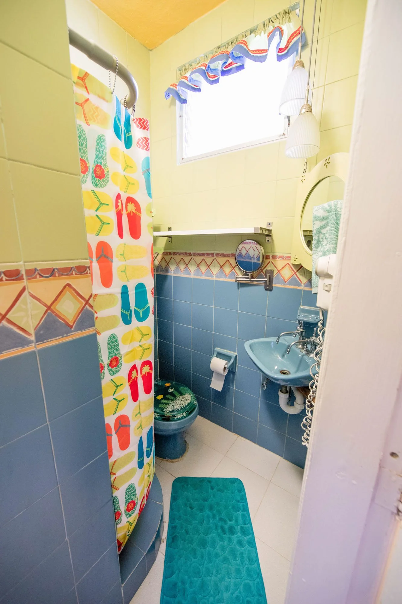 A small, colorful bathroom with a shower, teal toilet, small teal sink, and a bright blue bath mat. There is a window with a decorative valance, a mirror, and two hanging lamps.
