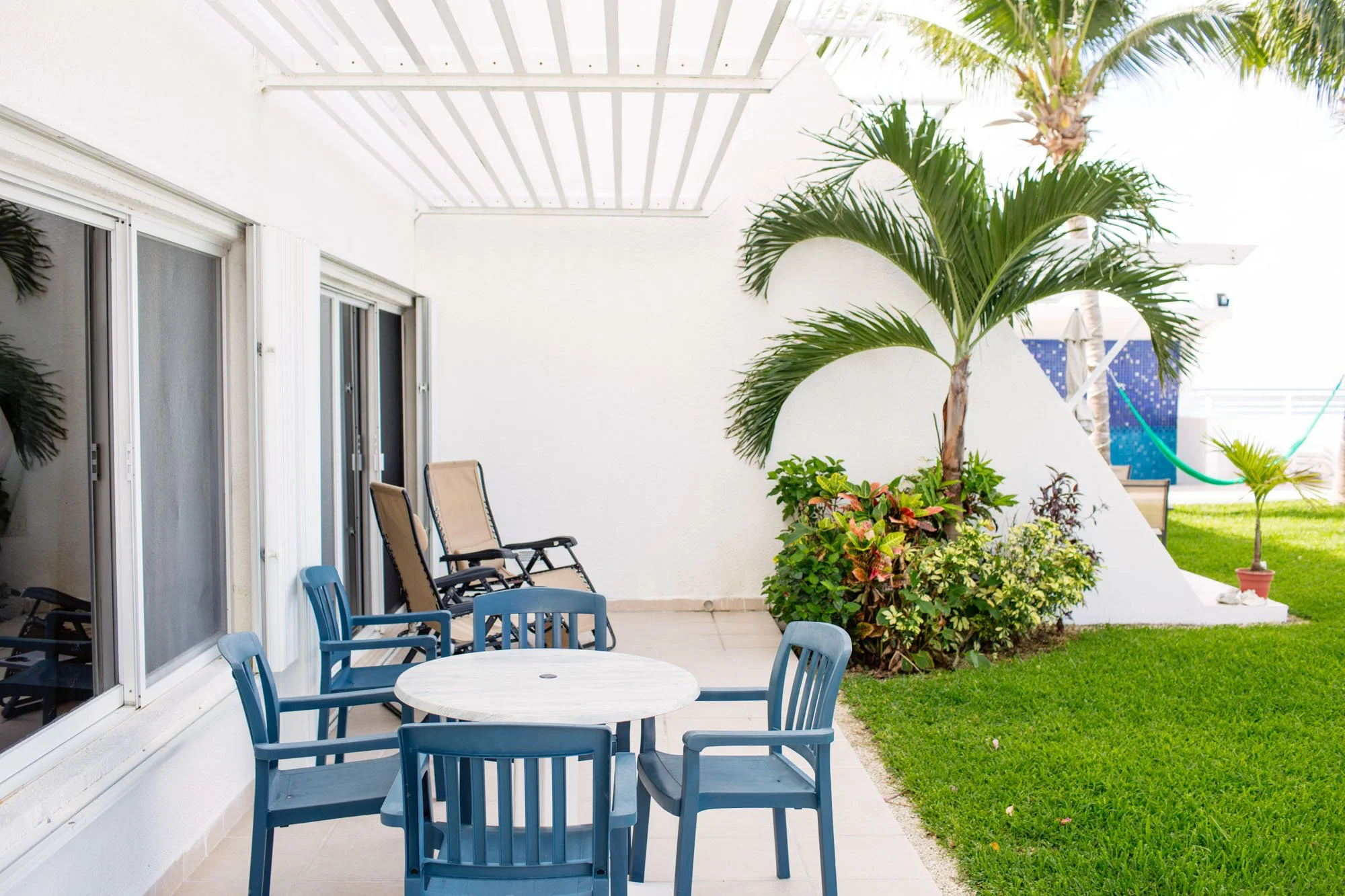 A patio at a Cozumel vacation rental with a round white table and six blue chairs, some patio lounge chairs, and a large palm tree with lush green plants nearby, garden, and grass area.