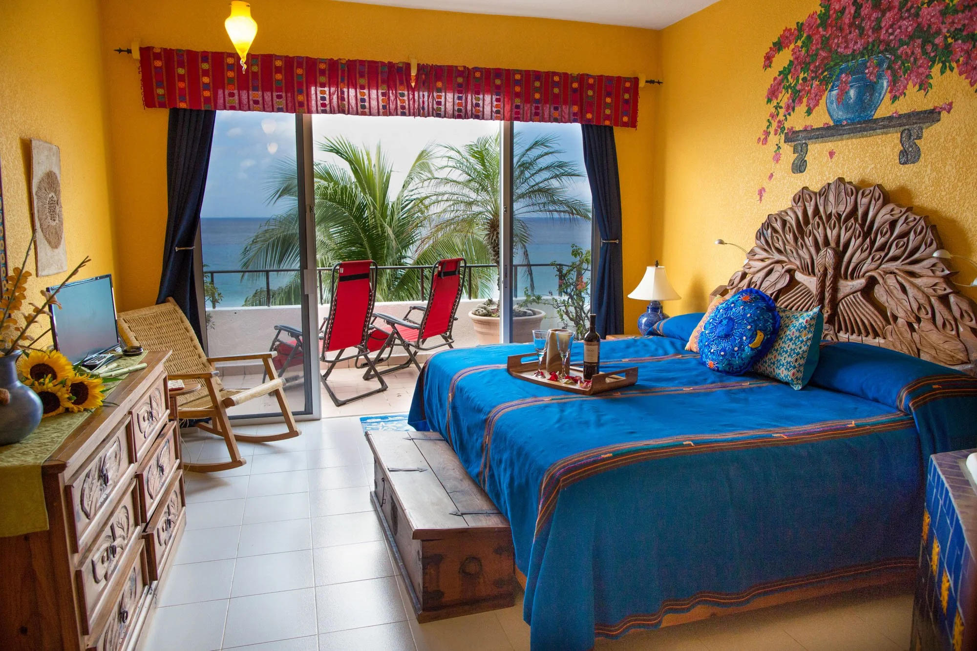 bed covered in blue linens, decorative pillows, and a carved wooden headboard. There is a tray with a wine bottle and glasses on the bed. A nightstand with a lamp is beside the bed. An open sliding door reveals a balcony with two red lounge chairs an