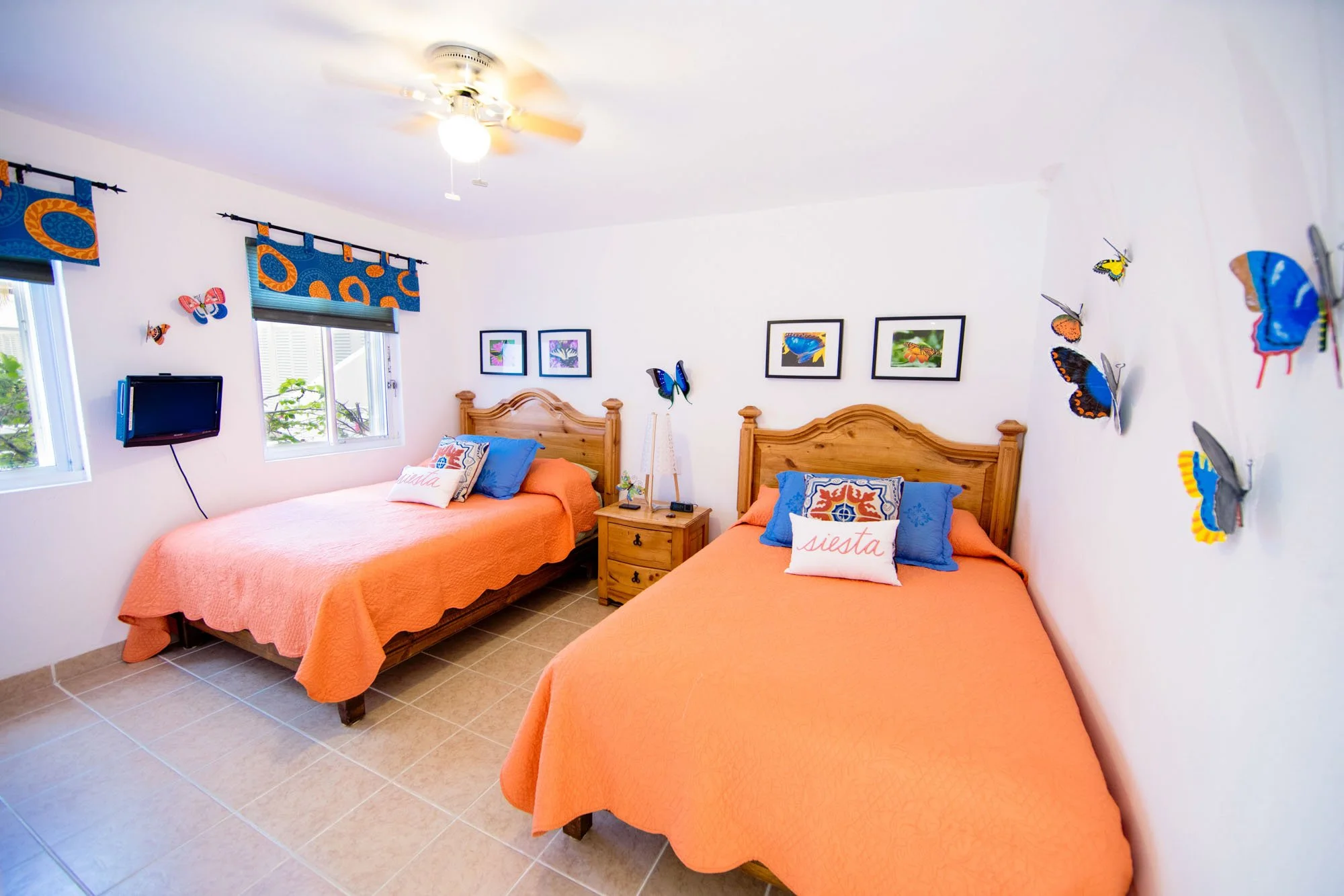 A bedroom with two twin beds with orange bedspreads, wooden headboards, and decorative pillows that say "siesta." There is a nightstand between the beds with a lamp, picture frames, and a small plant. The wall behind the beds has four framed pictures