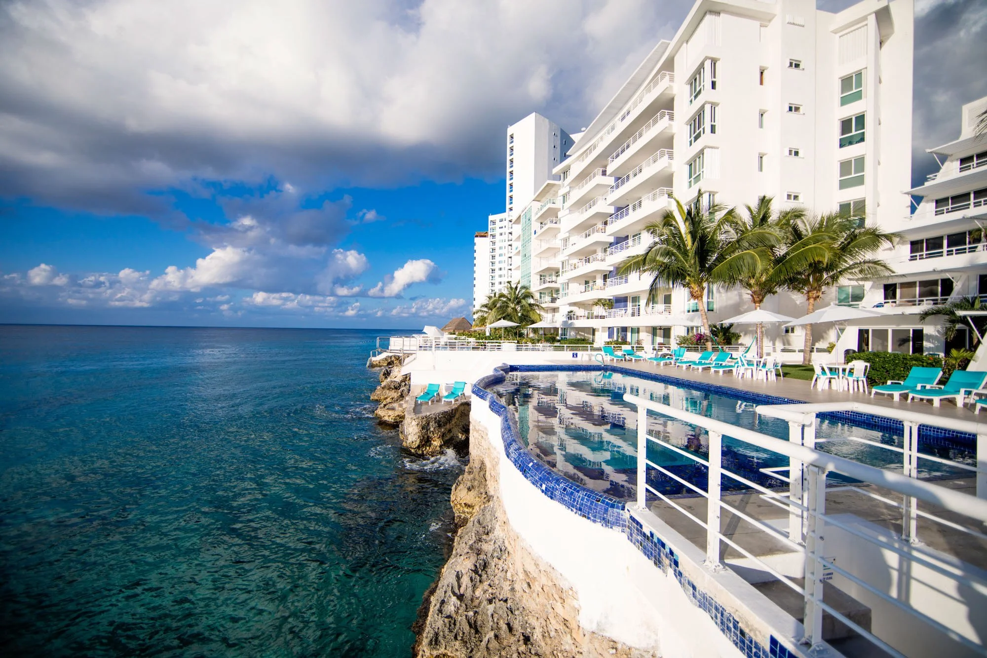 View Cozumel vacation rental by the ocean with a pool, white building, palm trees, and lounge chairs, under a partly cloudy sky.