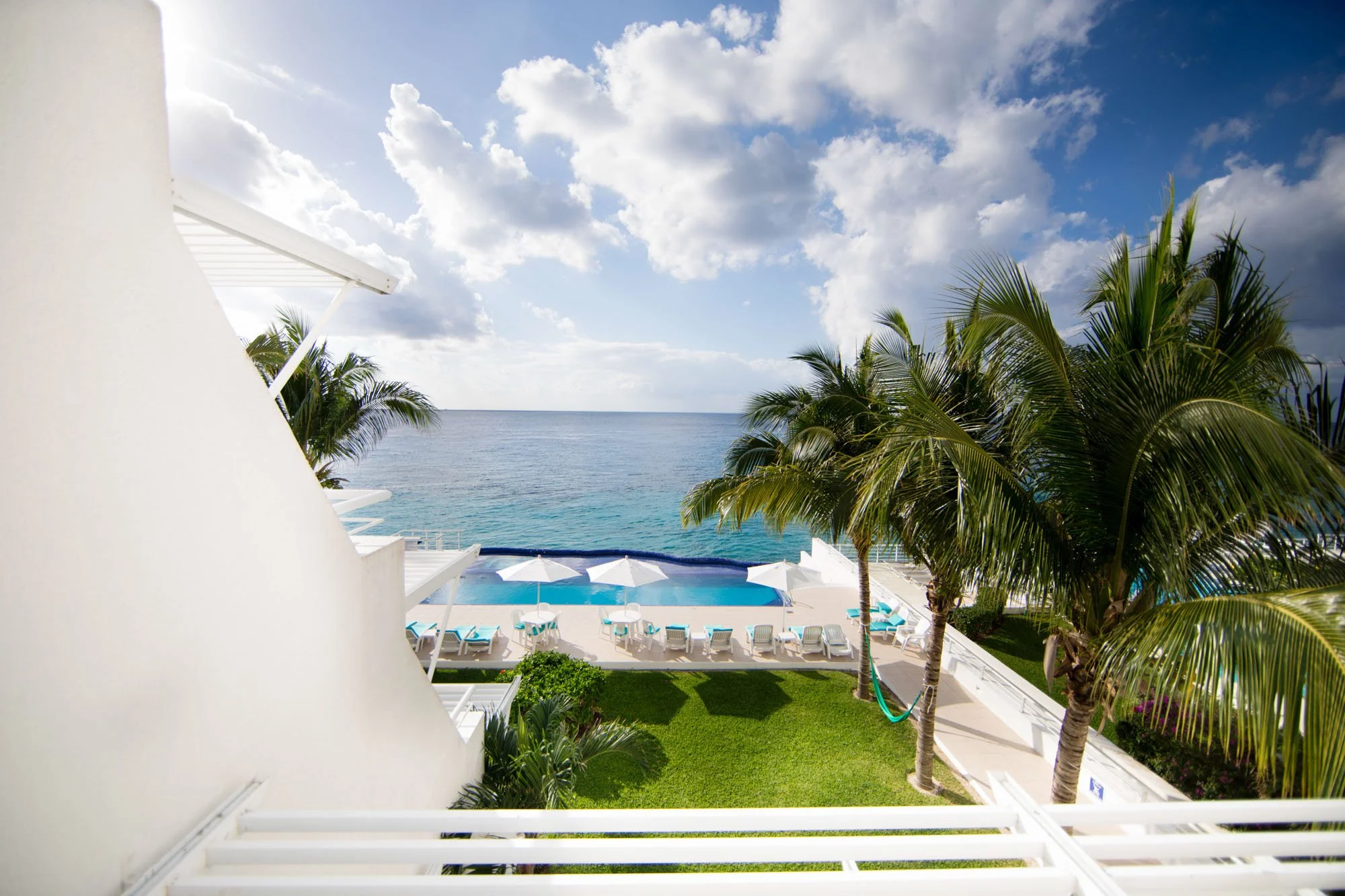 View of a tropical hotel with a swimming pool, white umbrellas, lounge chairs, lush green trees, and the ocean in the background under a partly cloudy sky.