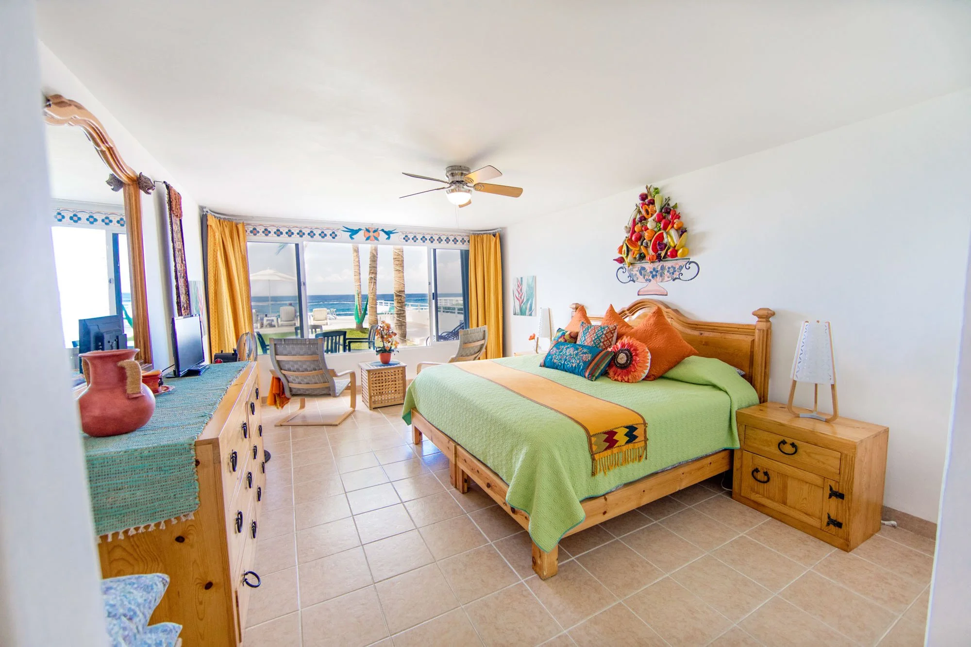 Beachside hotel room with a large bed, colorful pillows, and a wall-mounted fruit basket decoration. Has a ceiling fan, a dresser, a TV, a seating area near a large window with ocean view, and tropical curtains.