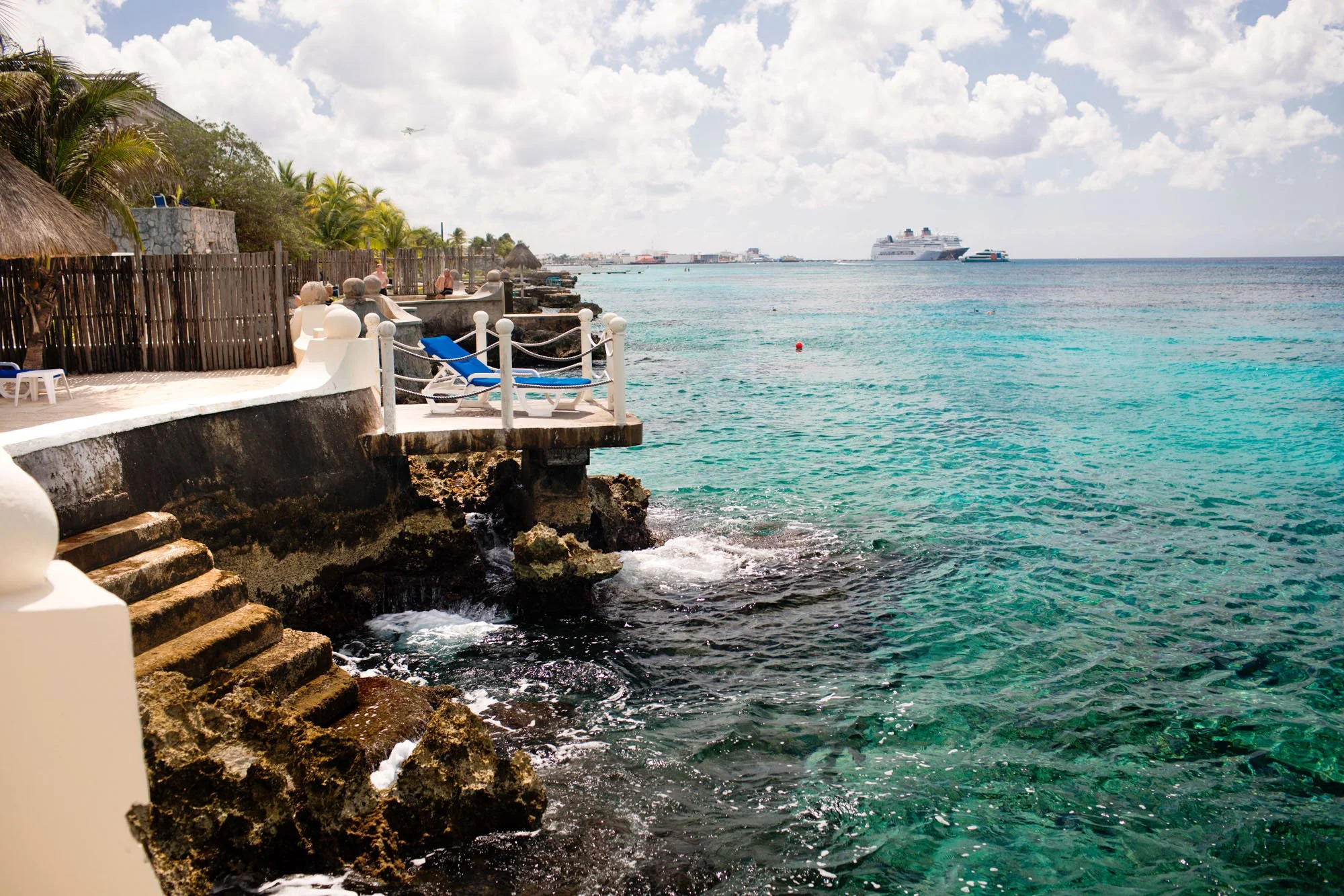 Seaside with steps leading to turquoise water, lounge chairs, palm trees, a cruise ship in the distance, and a partly cloudy sky.