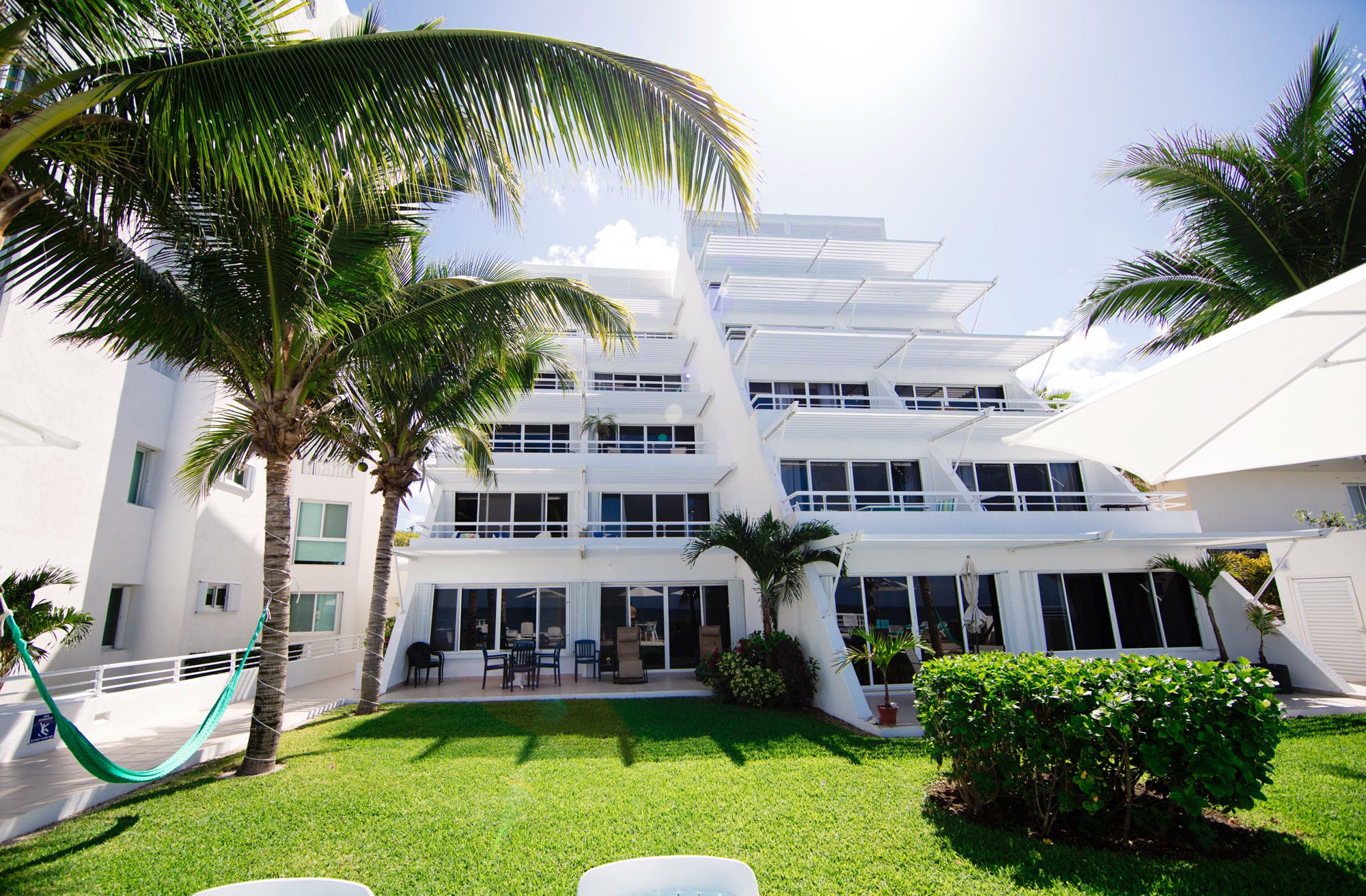 A white Cozumel vacation rental with balconies, surrounded by palm trees, with a lush green lawn in the foreground and outdoor seating area.