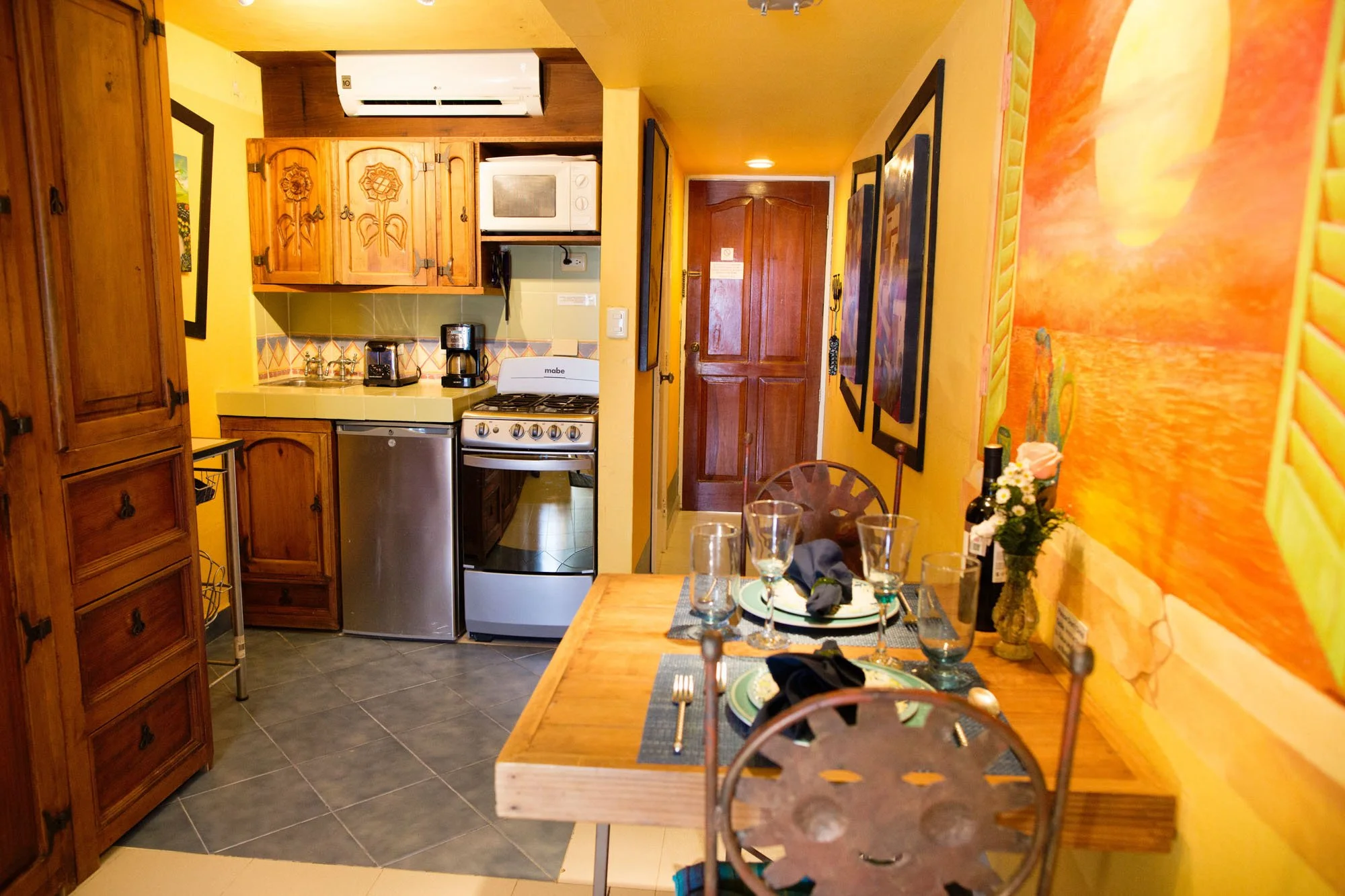 A cozy kitchen and dining area with yellow walls, wooden furniture, and artwork, set for a meal with glasses, plates, and black napkins on the dining table at a Cozumel vacation rental.