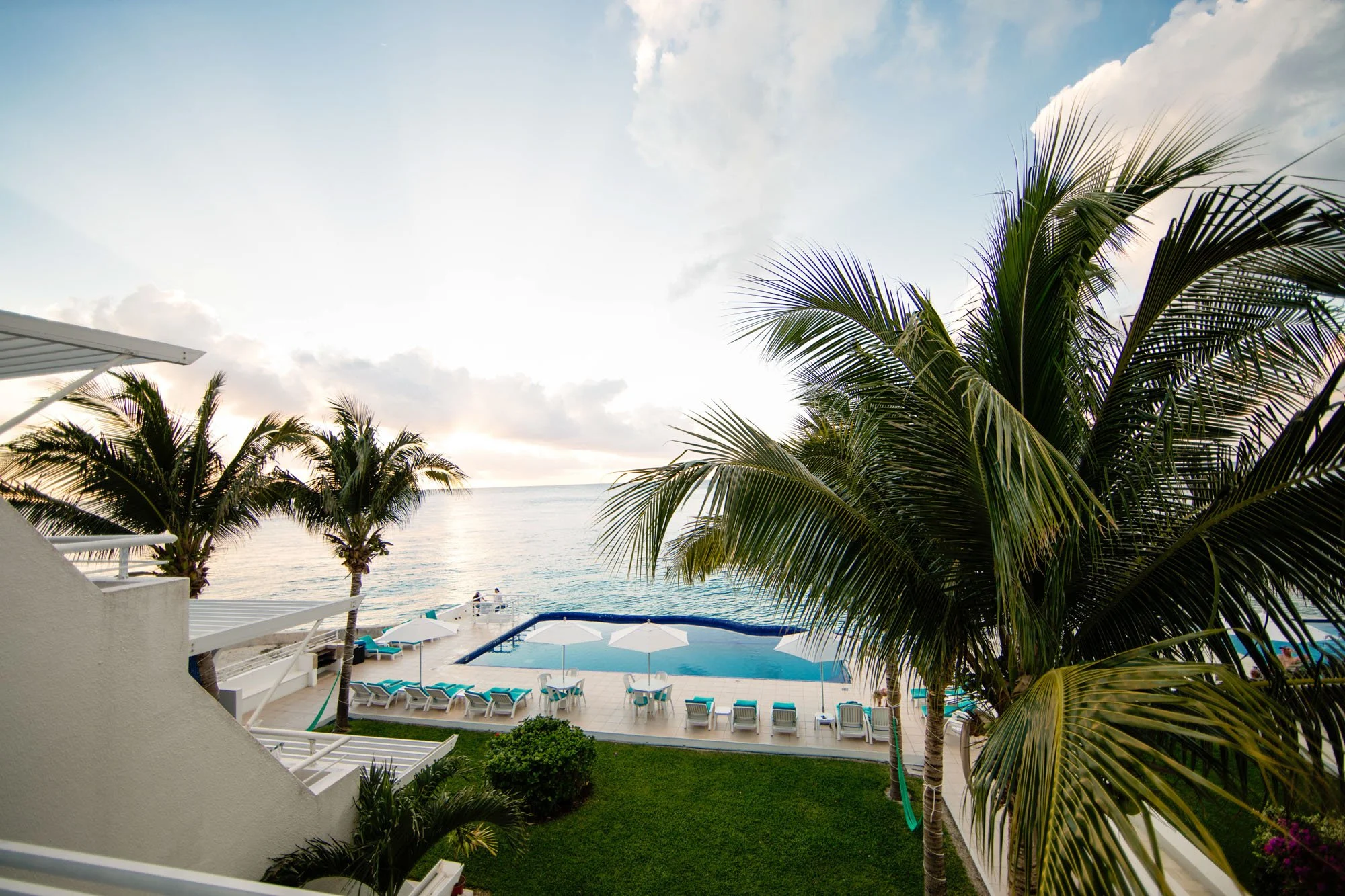 A view of a beachfront swimming pool surrounded by lounge chairs and umbrellas, with palm trees and the ocean in the background, under a partly cloudy sky.