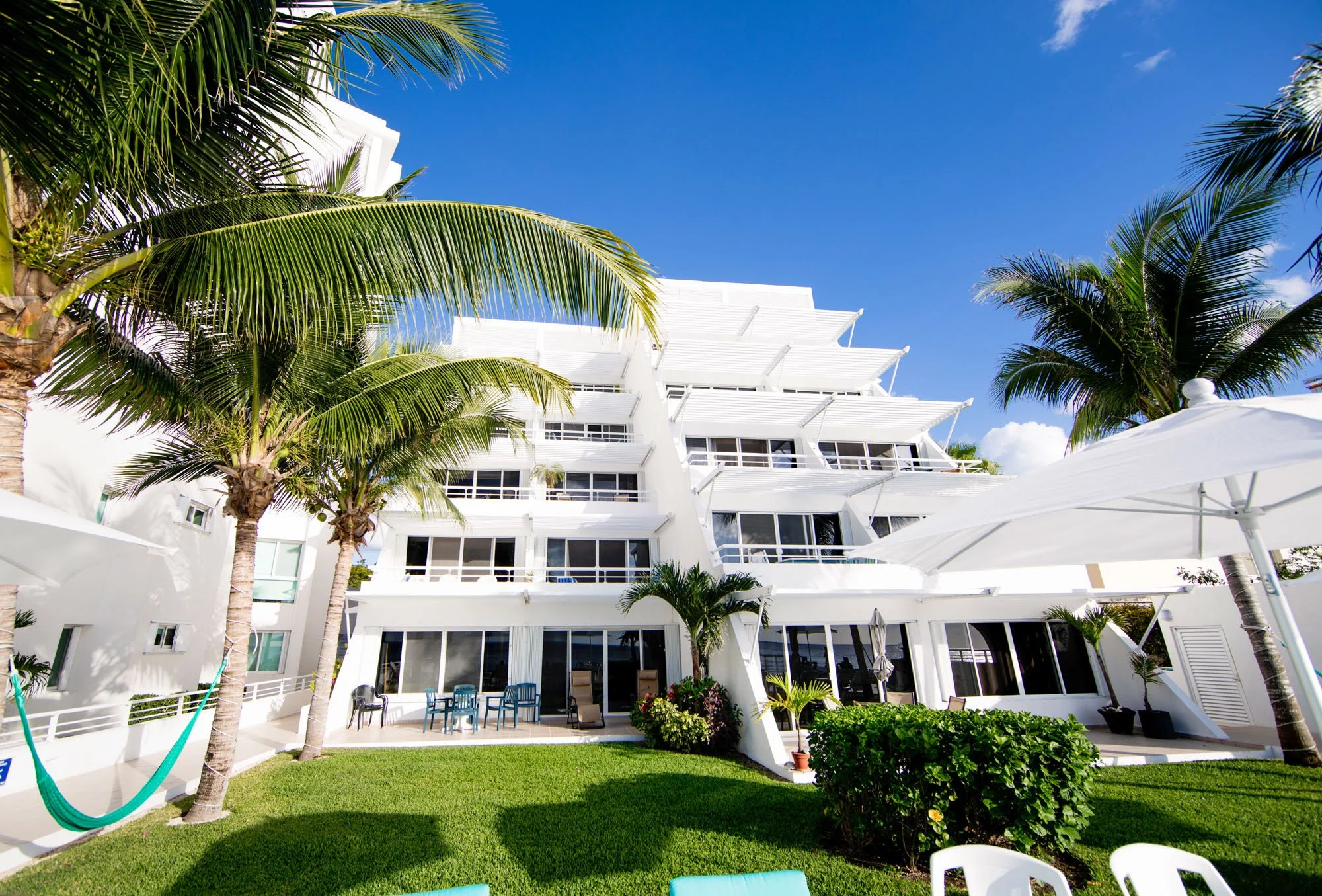 White multi-story hotel building with balconies, surrounded by palm trees and outdoor furniture, under a clear blue sky.