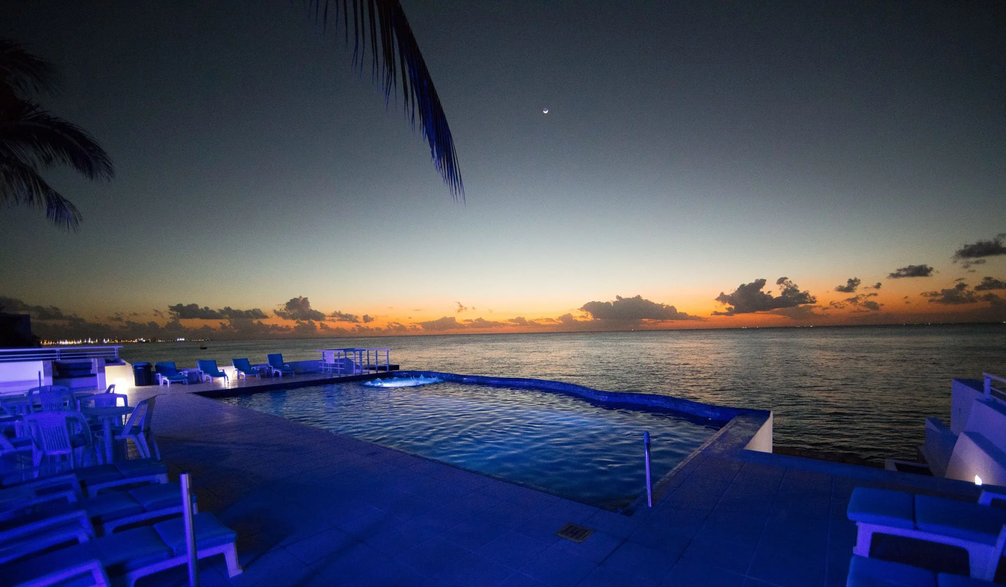 View of an infinity pool overlooking the ocean at sunset, with lounge chairs, a palm tree, and a crescent moon in the sky.
