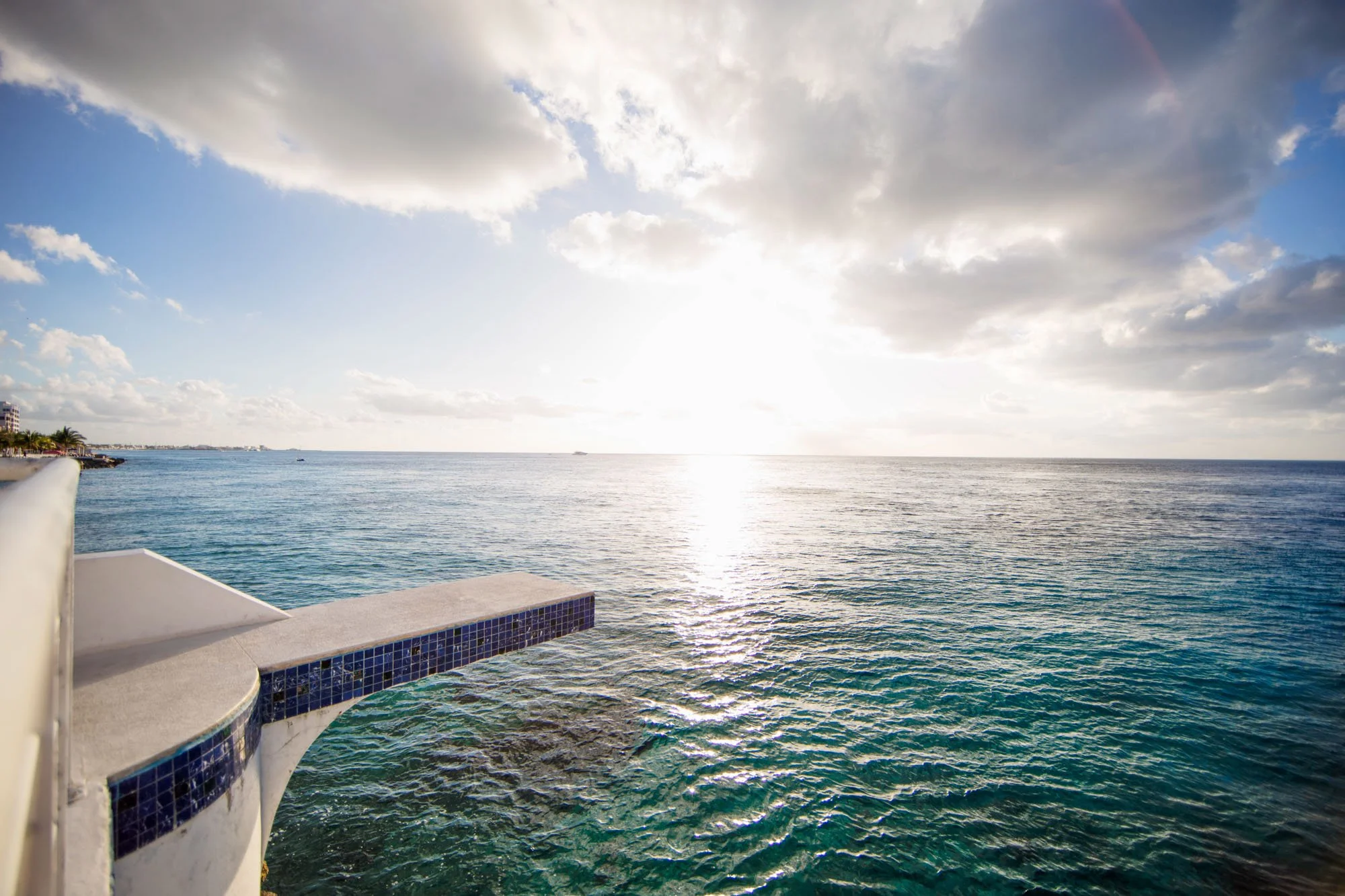 View of the ocean with sunlight reflecting on the water, clouds in the sky, and part of a building extending over the water.