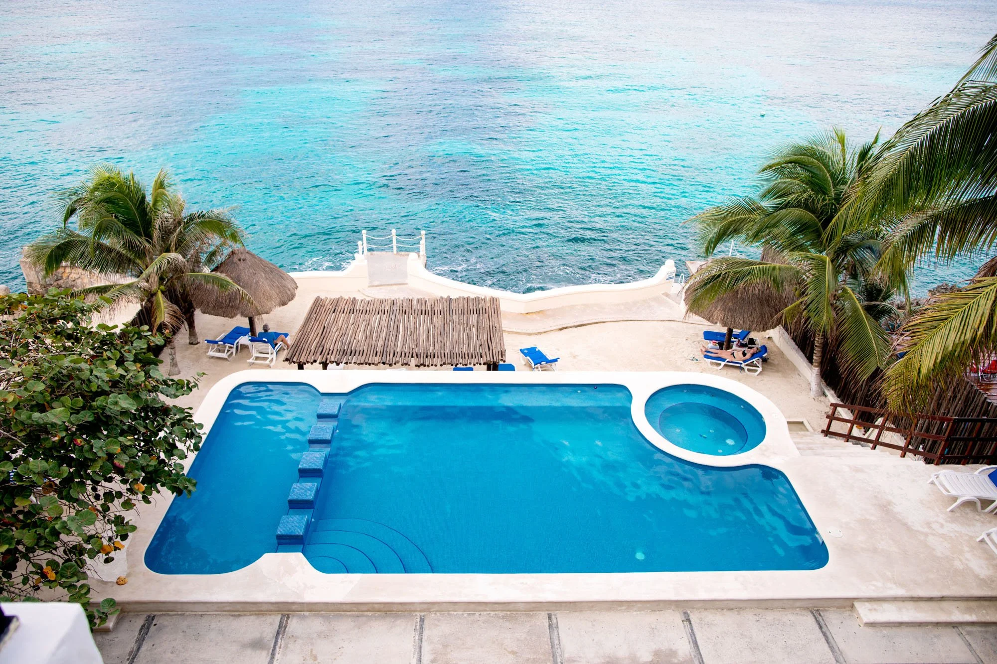 Swimming pool with a hot tub overlooking the ocean, surrounded by palm trees and lounge chairs on a beachside terrace at a Cozumel vacation rental.