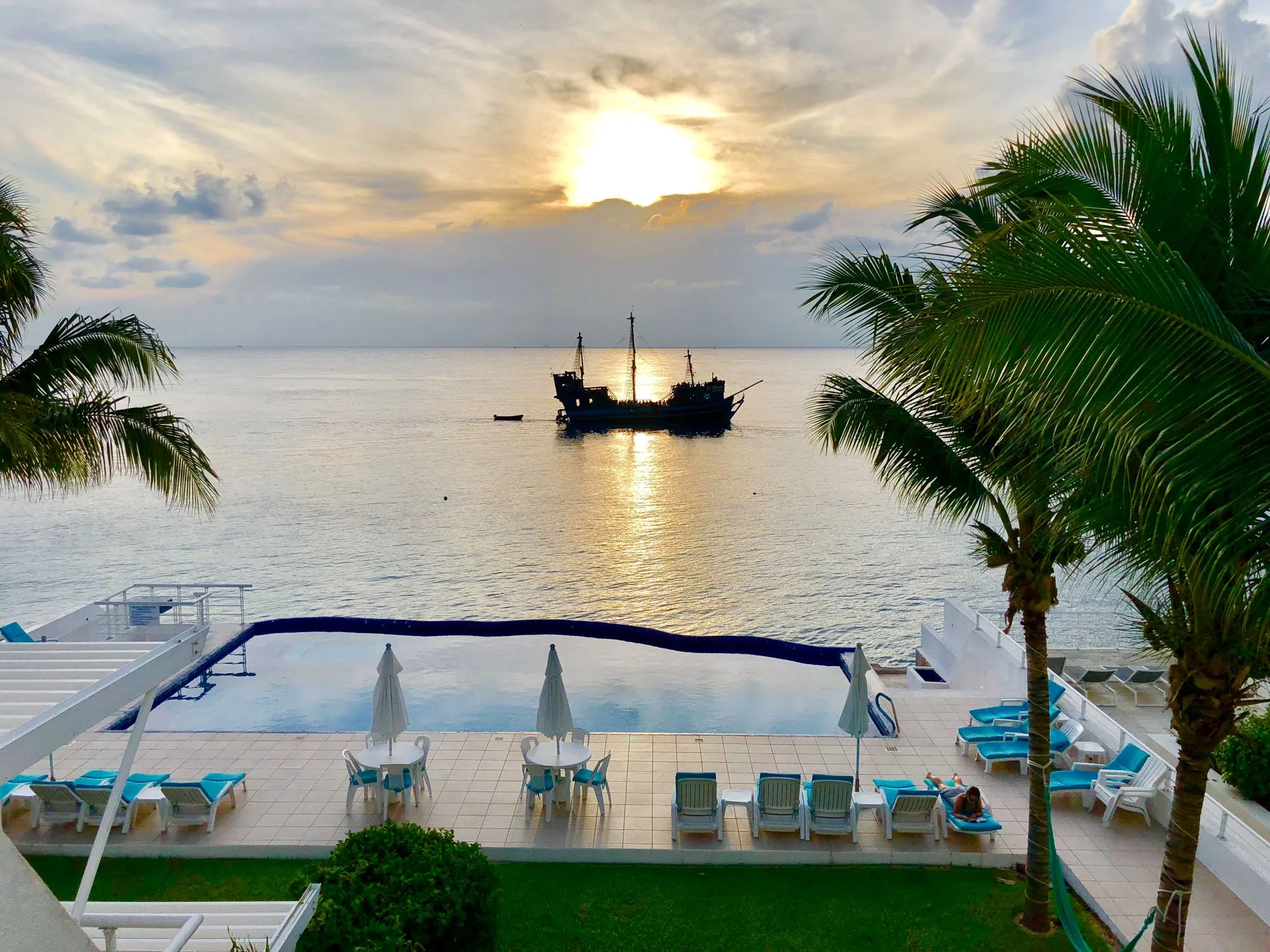 A sunset over a calm ocean with a pirate ship in the distance, viewed from a poolside area with lounge chairs and umbrellas, bordered by palm trees.