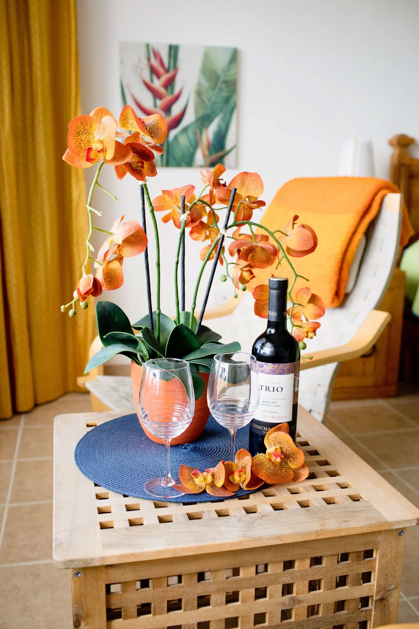 A table with a potted orange orchid, a bottle of red wine, two empty wine glasses, and orange flower petals. In the background, there is a white chair with an orange blanket, a framed botanical print, and yellow curtains.