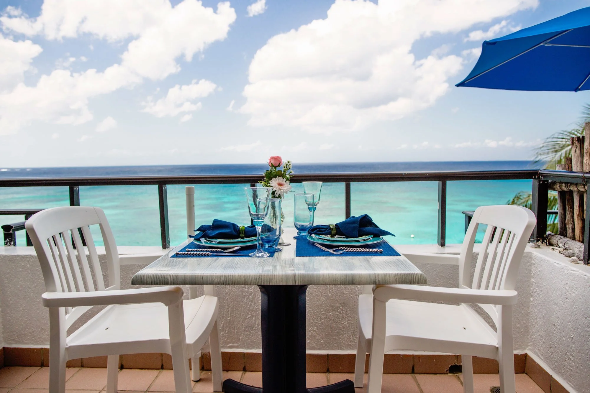 A seaside dining setup with a table set for two, decorated with a small flower vase, blue napkins, and glassware, overlooking turquoise ocean and a partly cloudy sky at a Cozumel vacation rental.