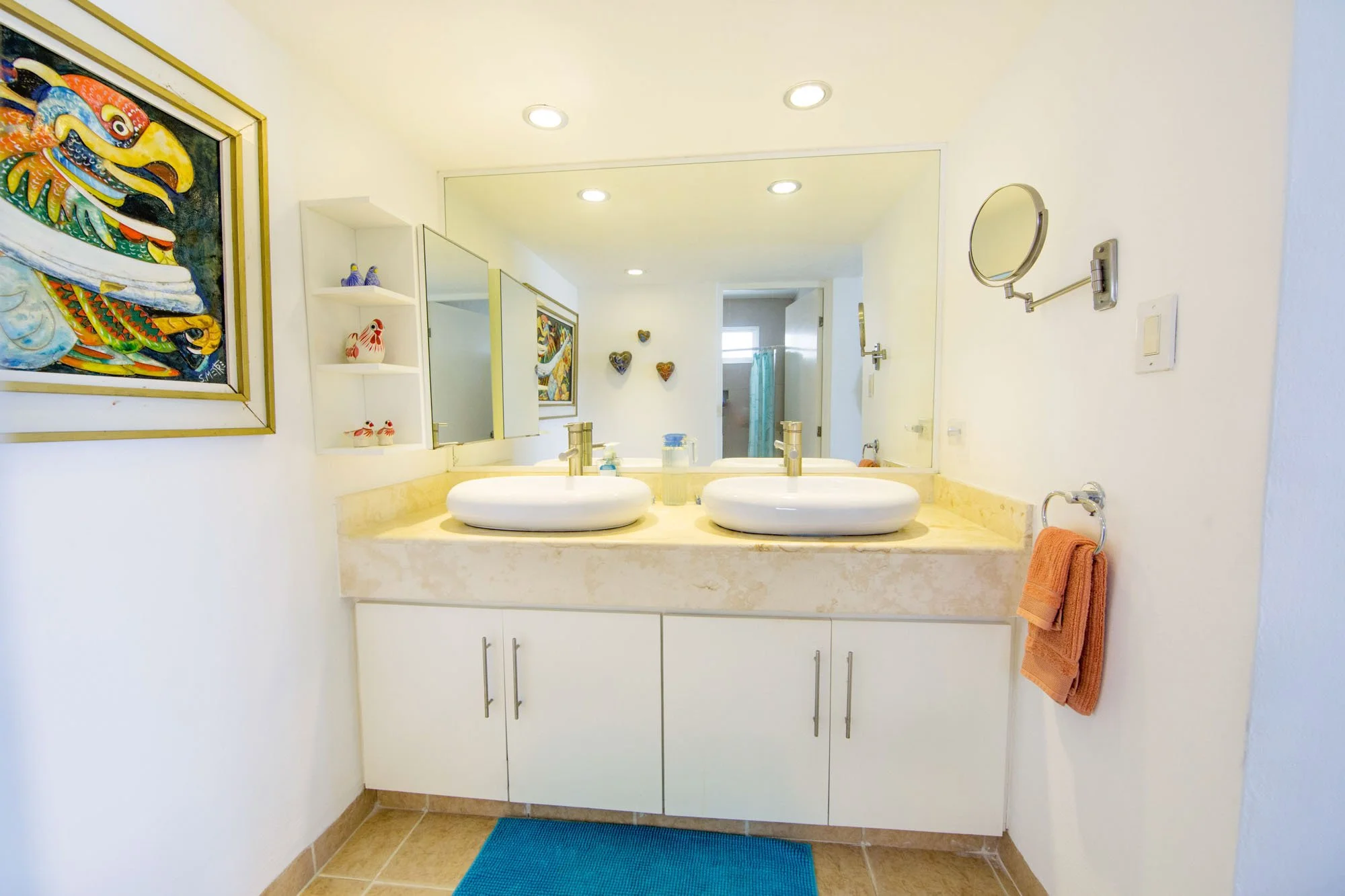Bathroom with double sinks, large mirror, wall art, towel rack with orange towels, and a colorful painting on the wall.