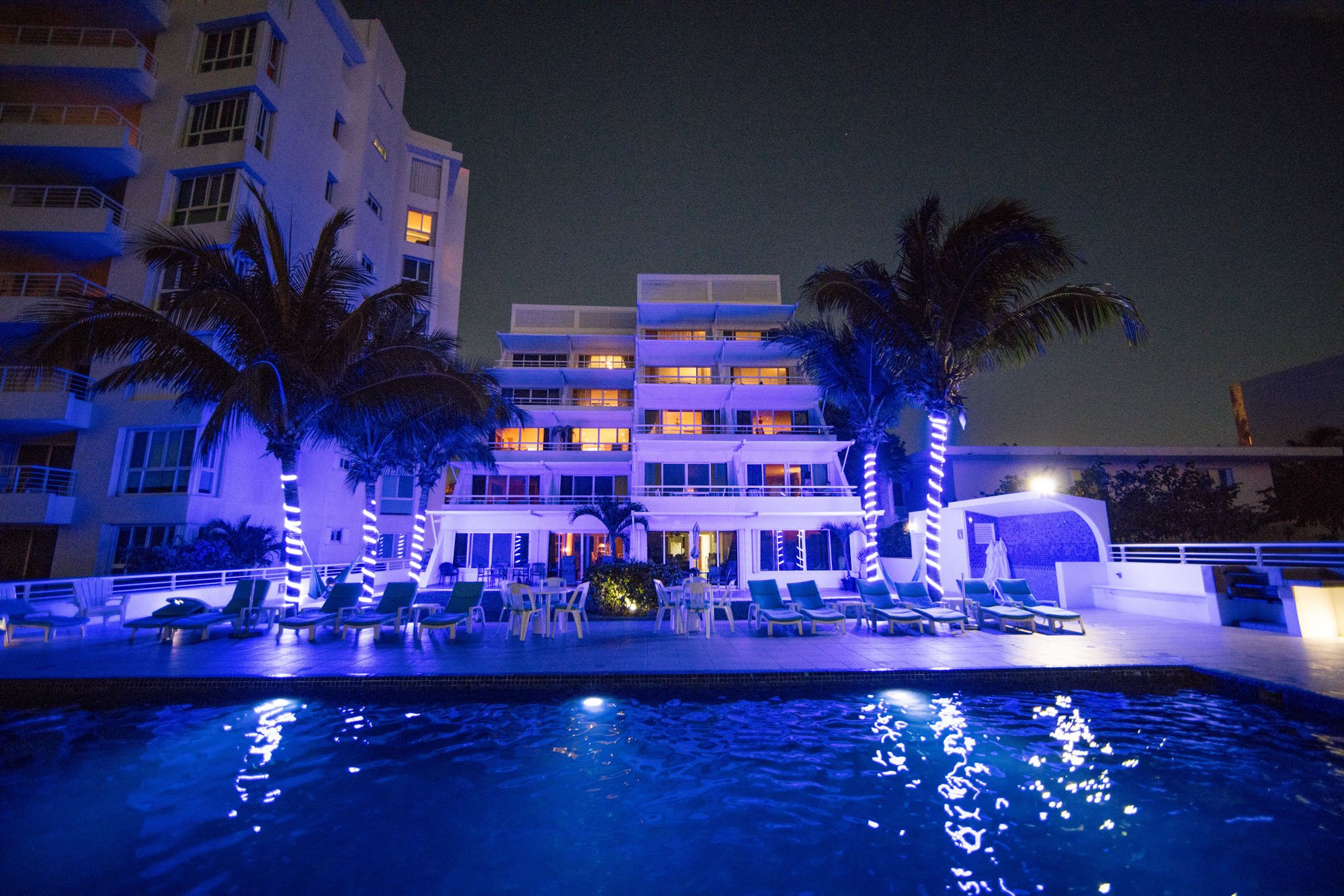 Nighttime view of a hotel with a swimming pool in the foreground, illuminated palm trees with blue and white lights, and the Cozumel vacation rental with lit windows in the background.