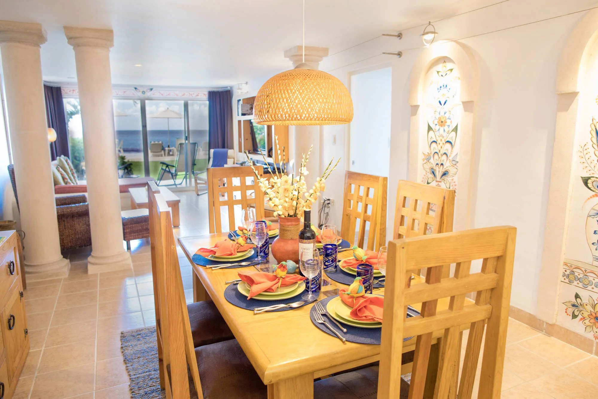Dining room with a wooden table set with colorful plates, napkins, glasses, and cutlery. A vase with white flowers is at center. In the background, there are sliding glass doors leading to a patio with outdoor furniture, and a view of the ocean.