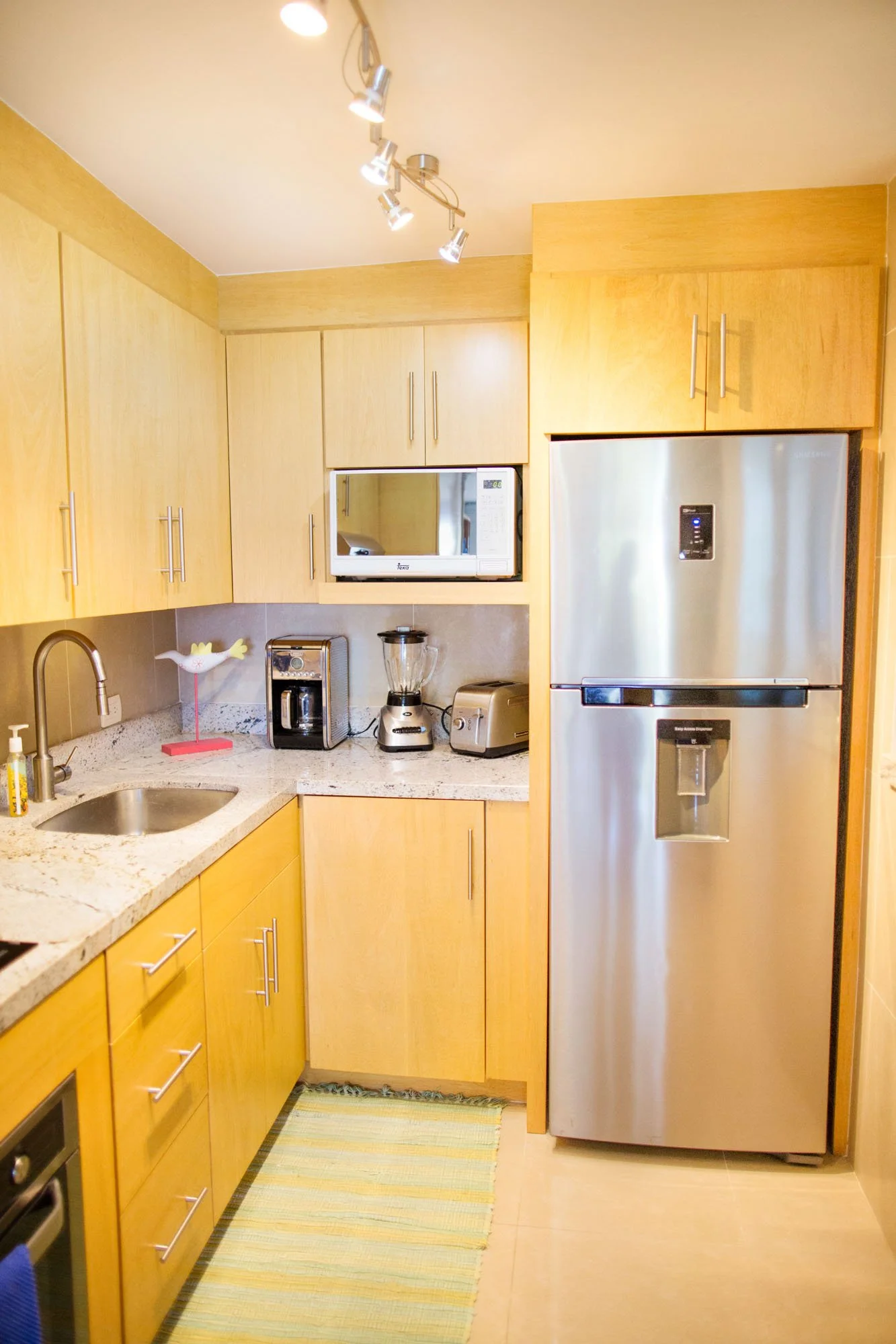 Compact kitchen with light wood cabinets, granite countertops, stainless steel refrigerator, microwave, coffee maker, blender, toaster, and small colorful striped rug.