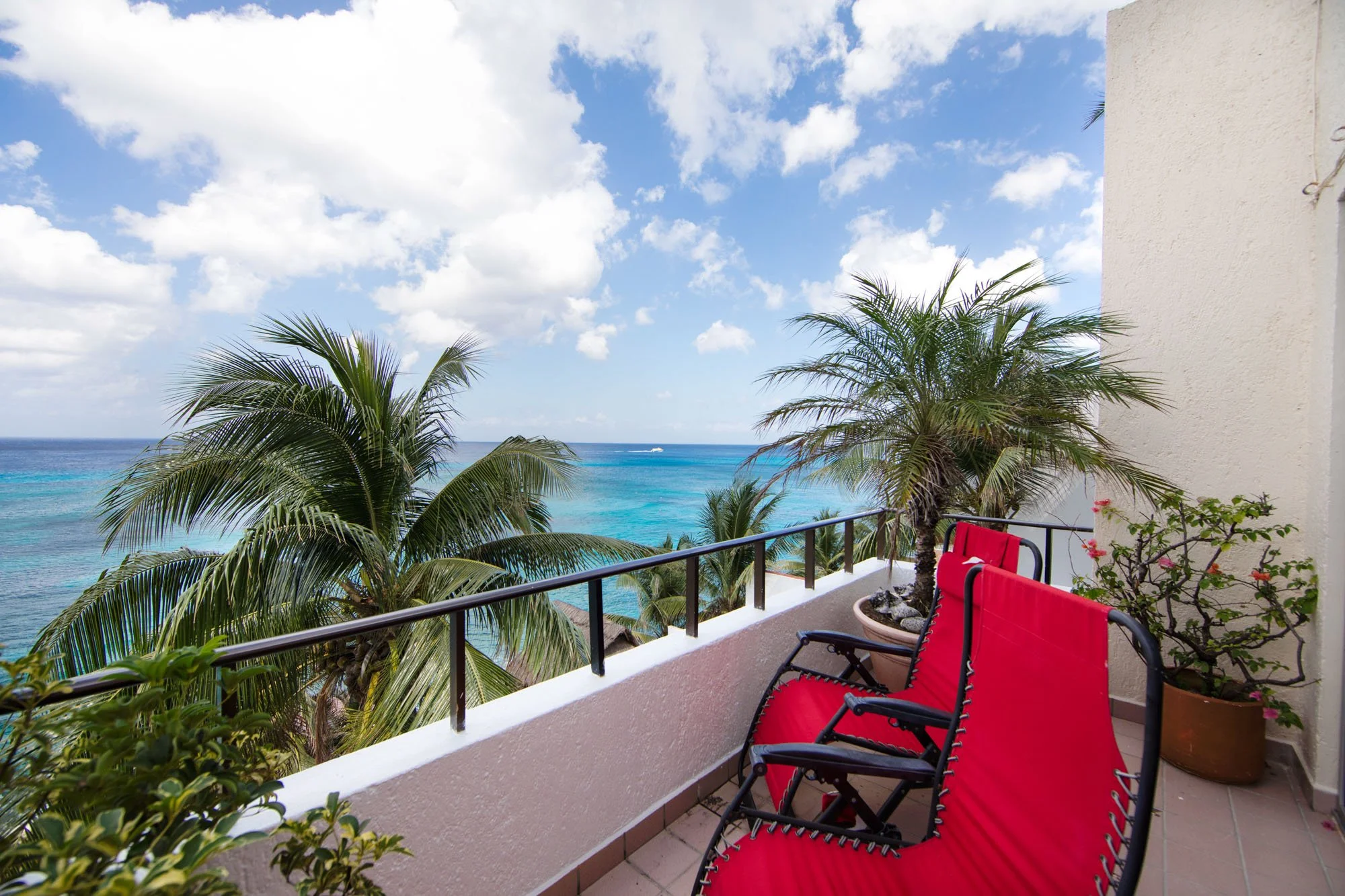 Balcony view with red lounge chairs, potted palm trees, and ocean scenery with palm trees, blue sky, and white clouds.