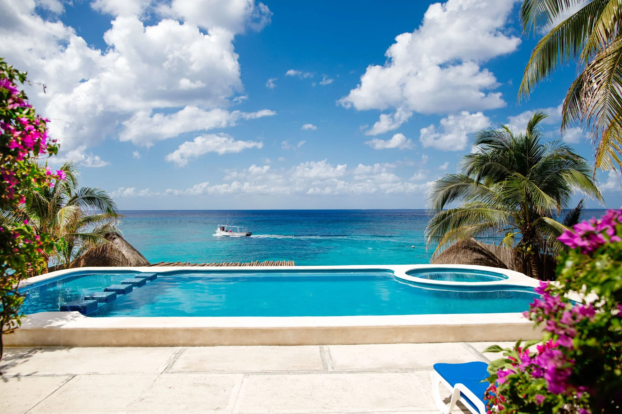 A tropical Cozumel vacation rental scene with a swimming pool overlooking the ocean, with a boat passing by, surrounded by palm trees and colorful flowers.