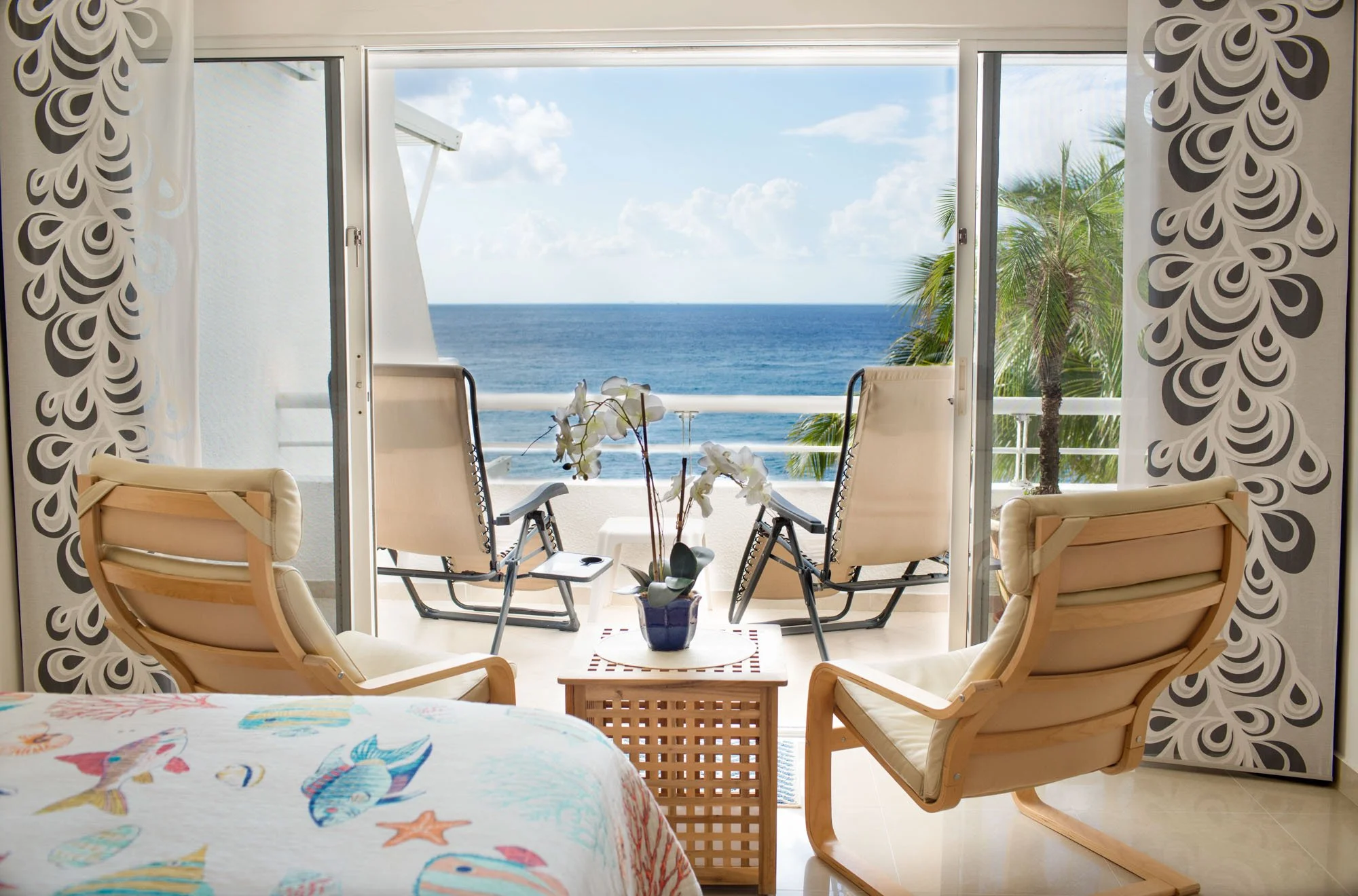A room with a bed, two comfortable chairs, and a small table with a potted orchid, overlooking a balcony with two chairs and an ocean view with palm trees.