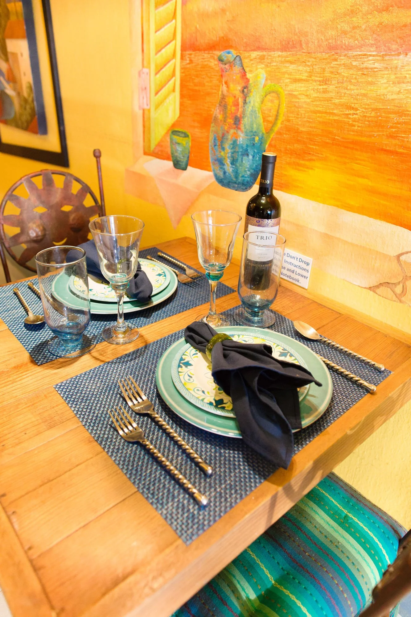 A table set for a meal with ceramic plates, silverware, napkins, wine glasses, and a bottle of wine in a bright dining area with colorful artwork on the wall at a Cozumel vacation rental.