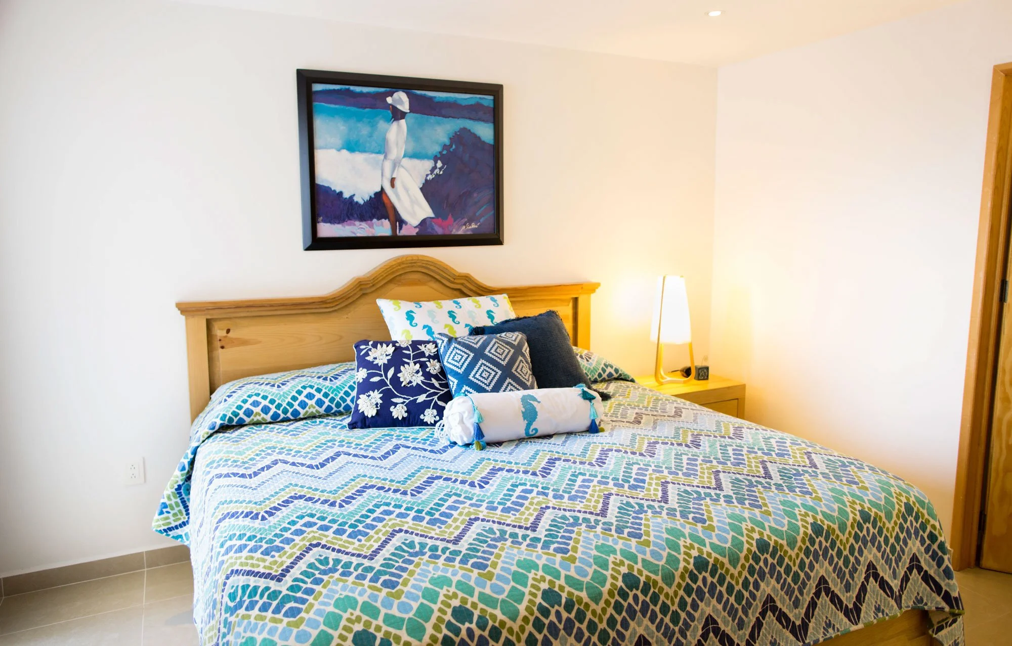 A bedroom with a wooden bedframe, patterned bedspread, and multiple decorative pillows. A nightstand with a lamp is beside the bed. A colorful painting of a woman in a white dress and hat hangs above the bed.