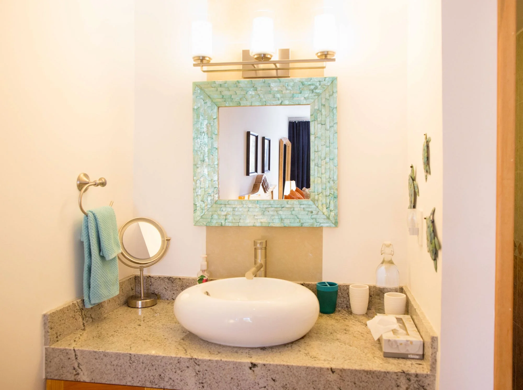 Bathroom vanity area with a white vessel sink, soap, a mirror, and decorative items. Towel hanging on a ring to the left, three wall-mounted candles above, and art on the wall to the right.