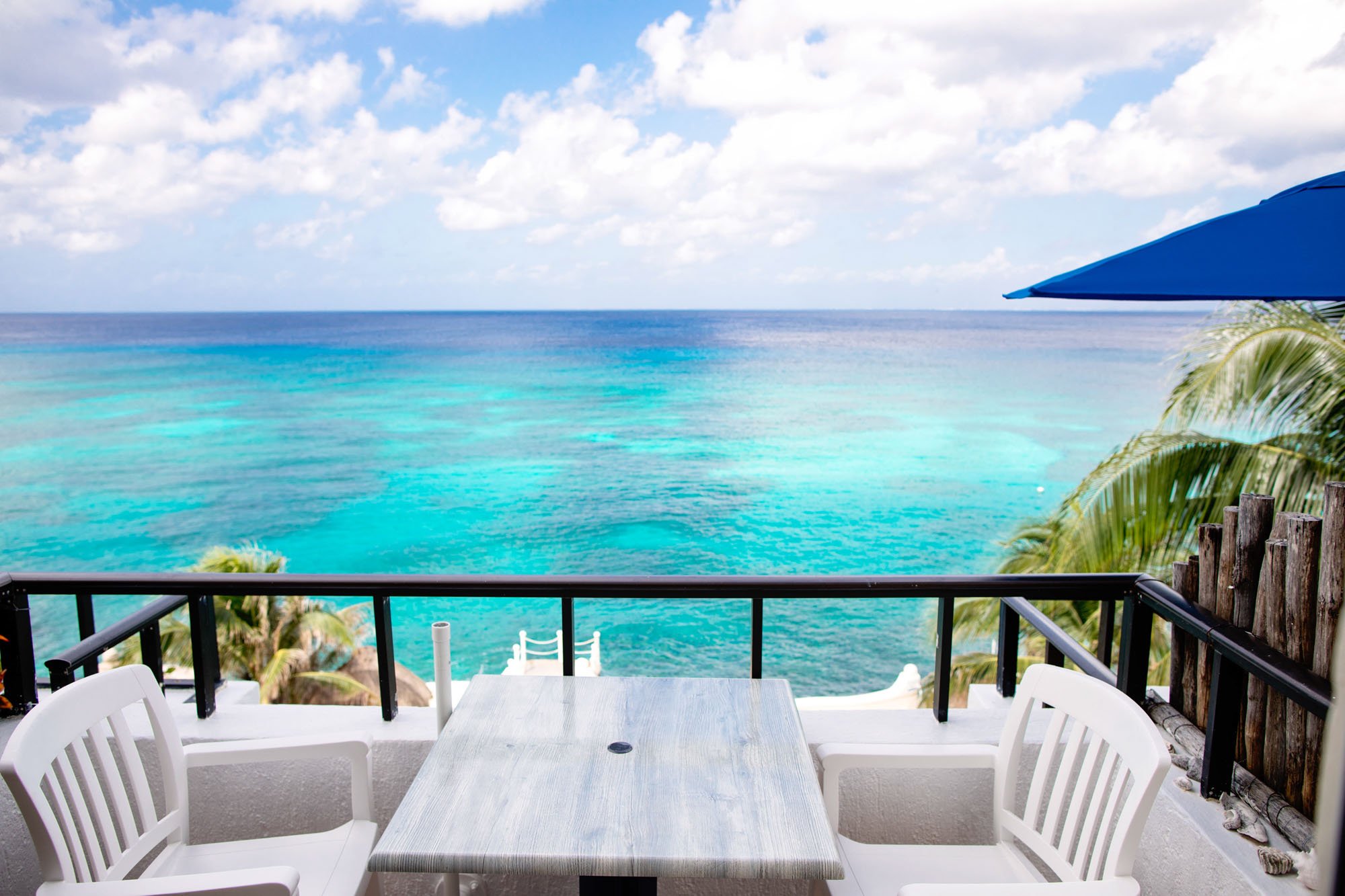 View of the ocean from a Cozumel vacation rental balcony with a table, chairs, an umbrella, and tropical palm trees.