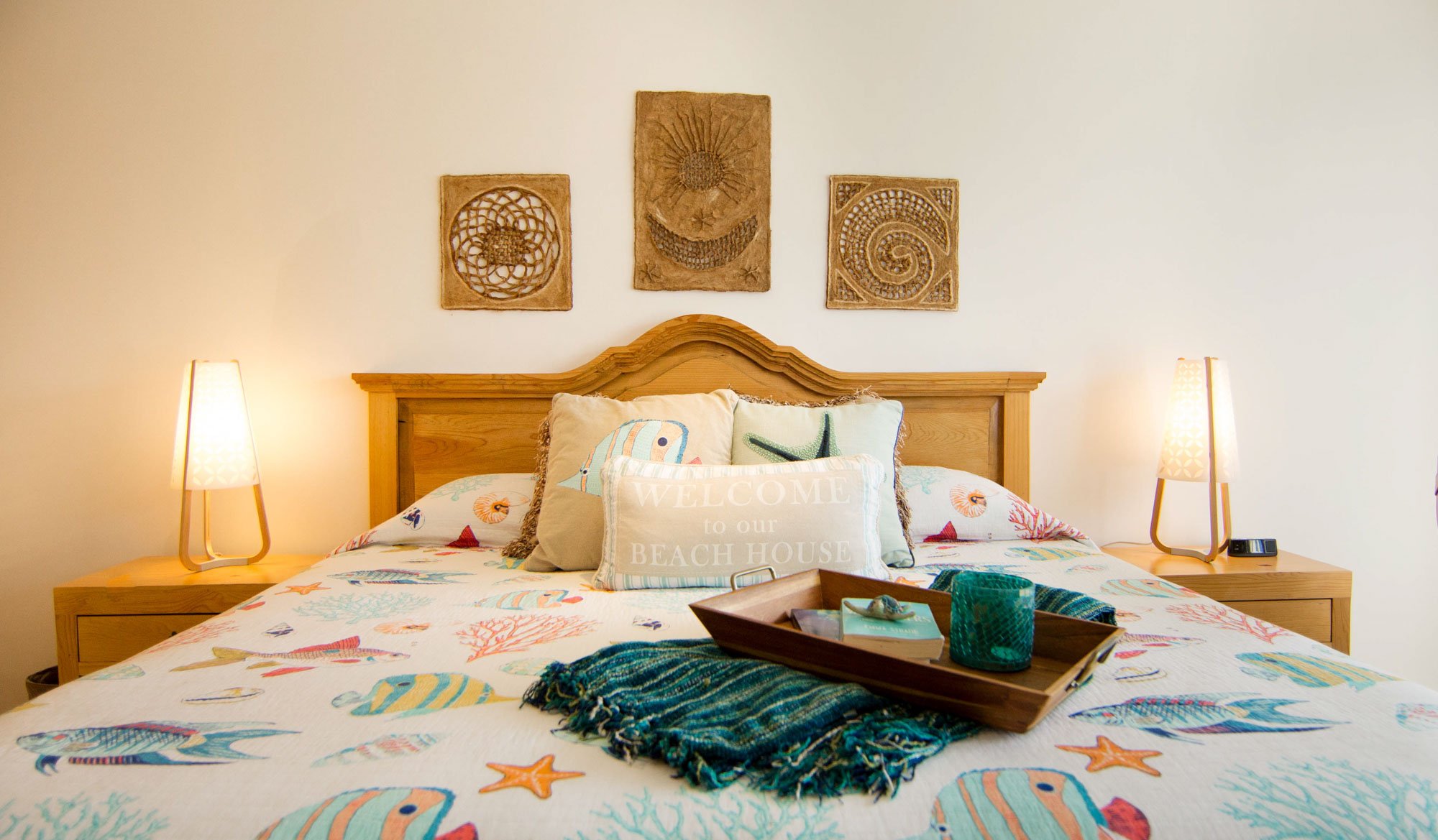 Beach-themed bedroom with wooden bed, colorful ocean animal bedding, decorative pillows, and wall art, illuminated by two bedside lamps.