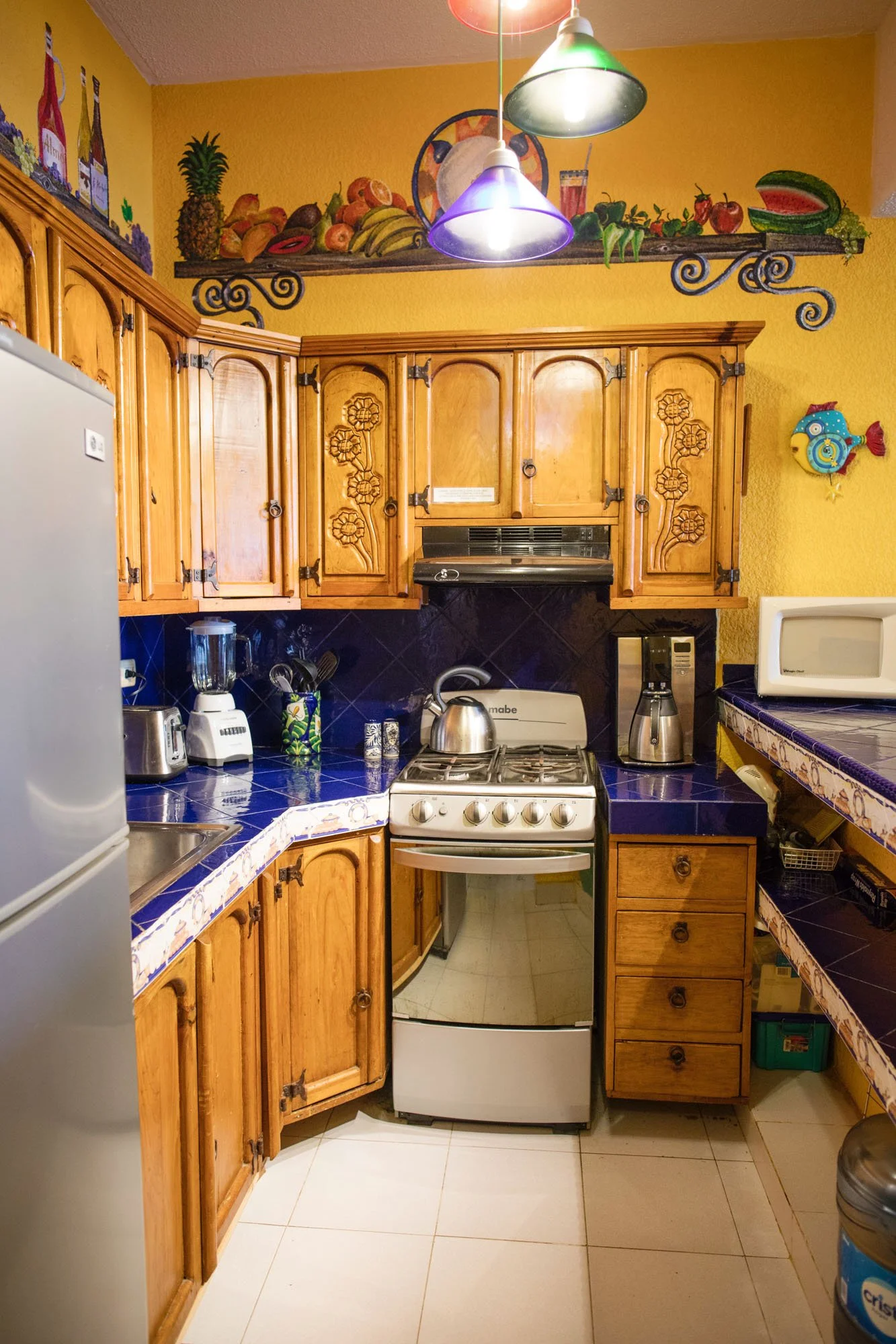 A kitchen with wooden cabinets, blue tiled countertops, and various appliances including a stove, microwave, toaster, blender, and coffee maker. There is a colorful fruit mural on the yellow wall above the cabinets and a decorative fish wall hanging 