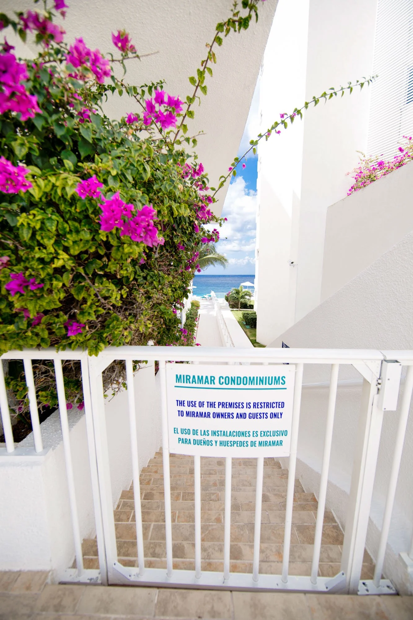 Gate with a sign in front of a building at Miramar Condominiums, with bougainvillea and a view of the ocean in the background.