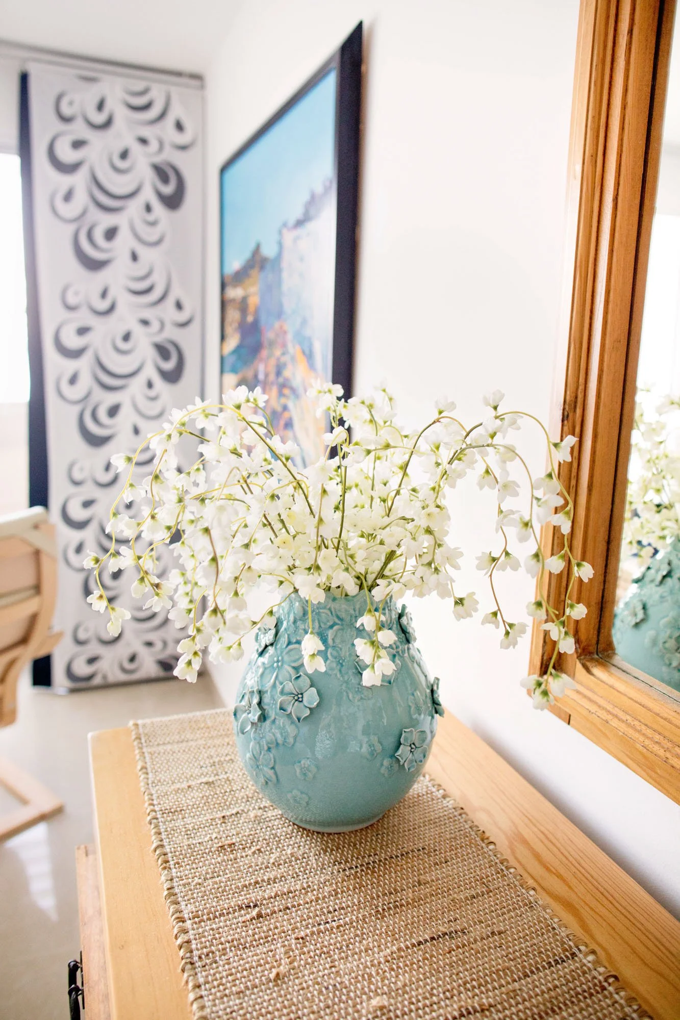 A blue vase with floral designs holding white paperwhite flowers on a wooden table with woven placemat, next to a mirror, with a framed painting in the background.