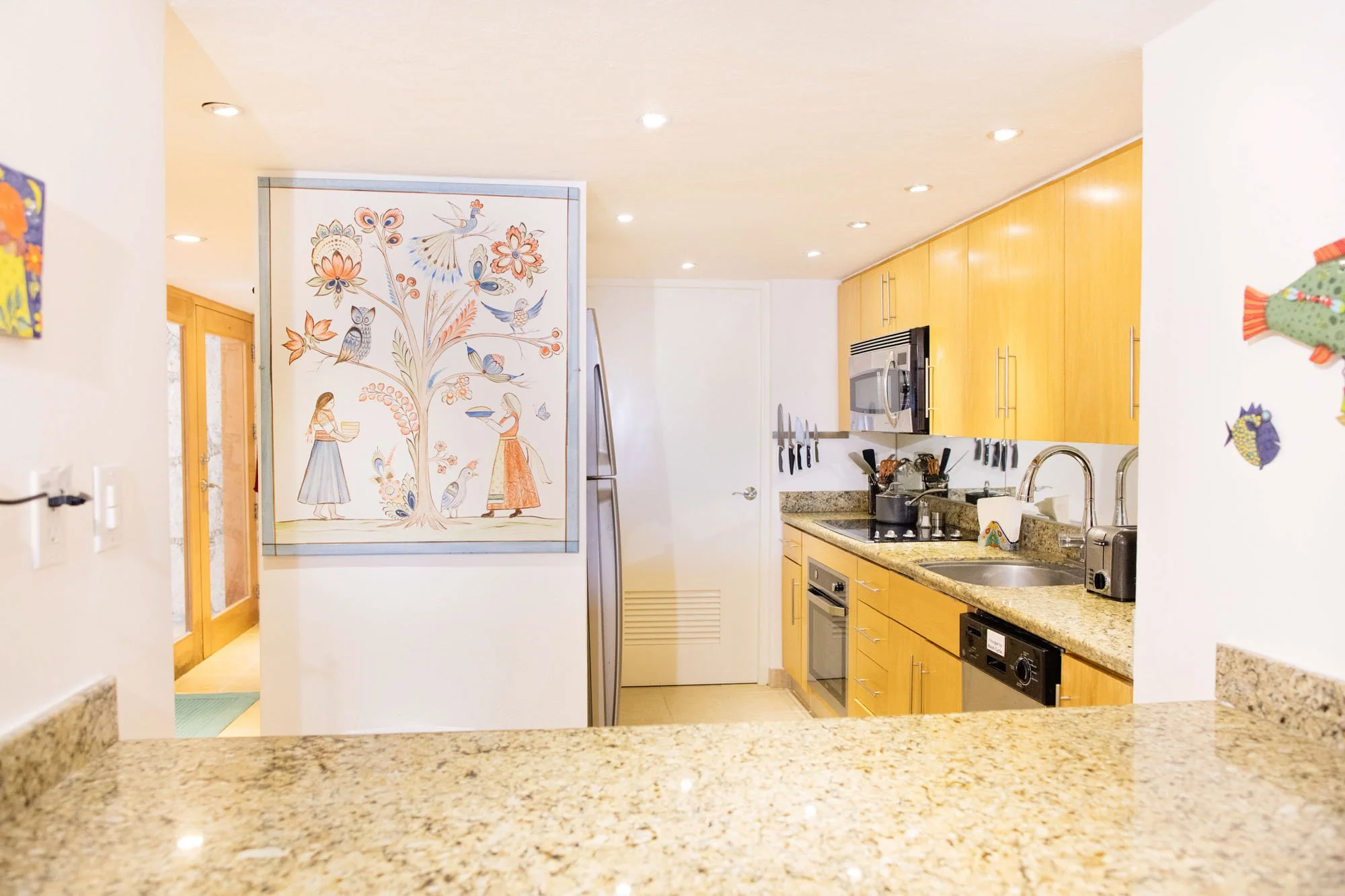 A modern kitchen with wooden cabinets, granite countertops, stainless steel appliances including microwave and oven, and various kitchen utensils. There is an art piece with colorful bird and floral motifs on the wall and a view into an entryway with
