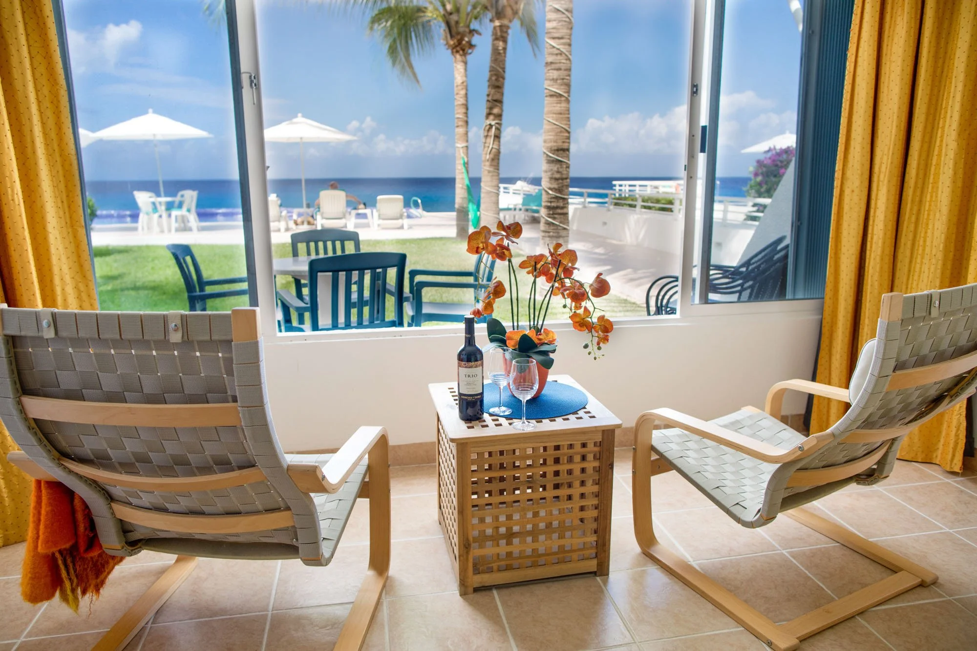 View through Cozumel vacation rental window showing a patio with lounge chairs, an ocean, blue sky with clouds, and palm trees.