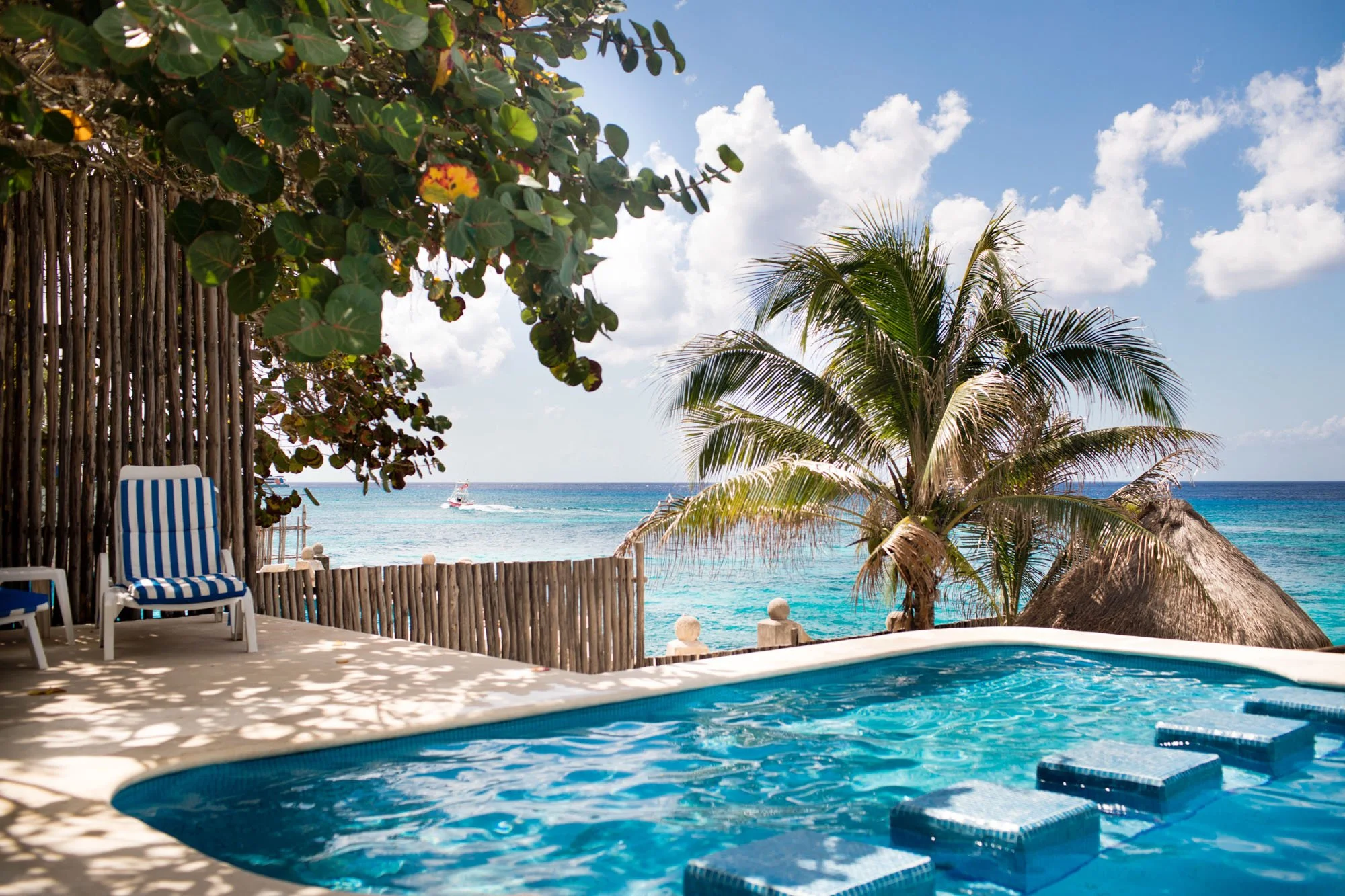 A swimming pool overlooking a beach with a palm tree and thatched roof, blue sky with clouds, and a boat in the water.