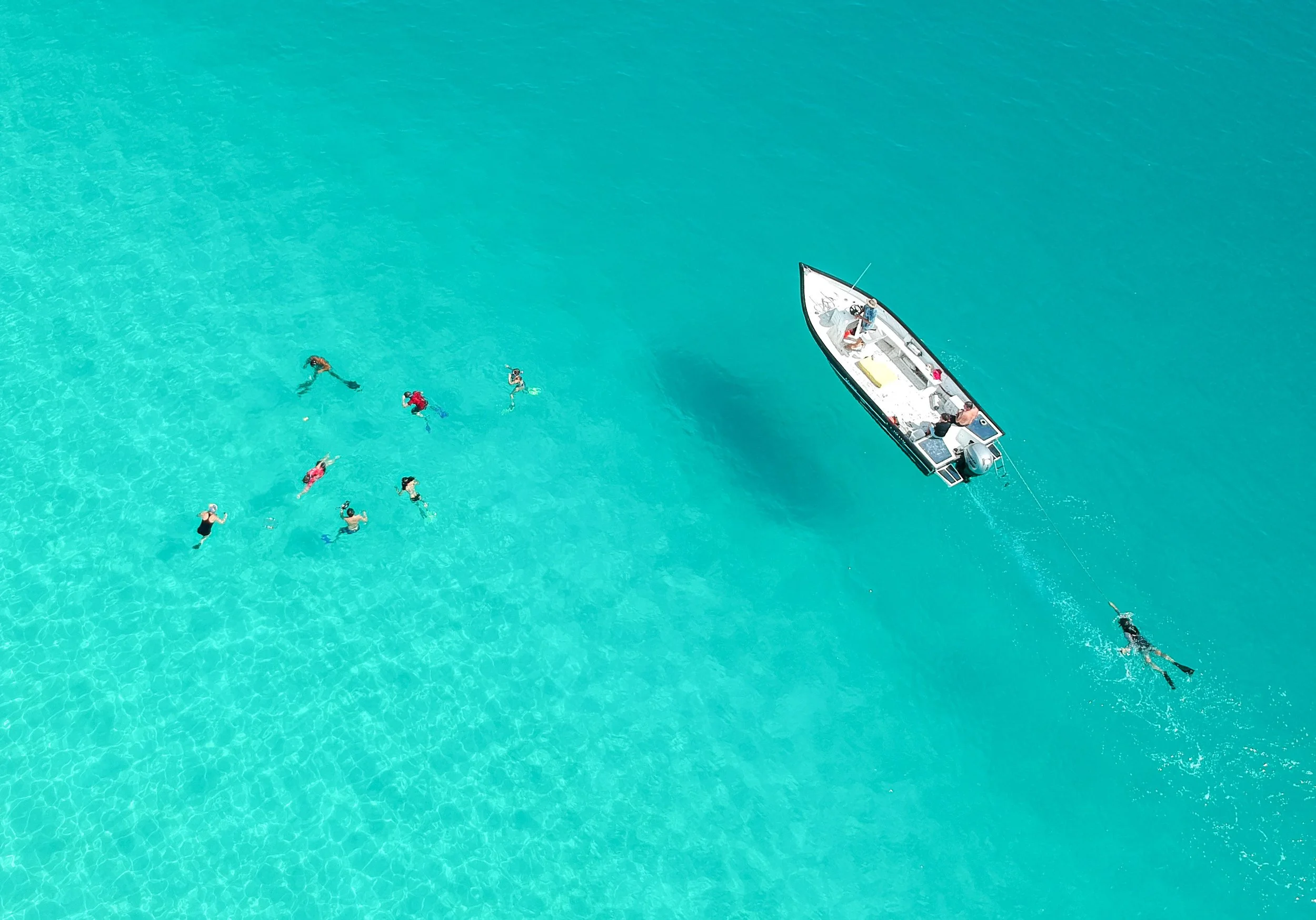 Aerial view of a group of people swimming in clear blue water near a boat, with one person being pulled on a line behind the boat.