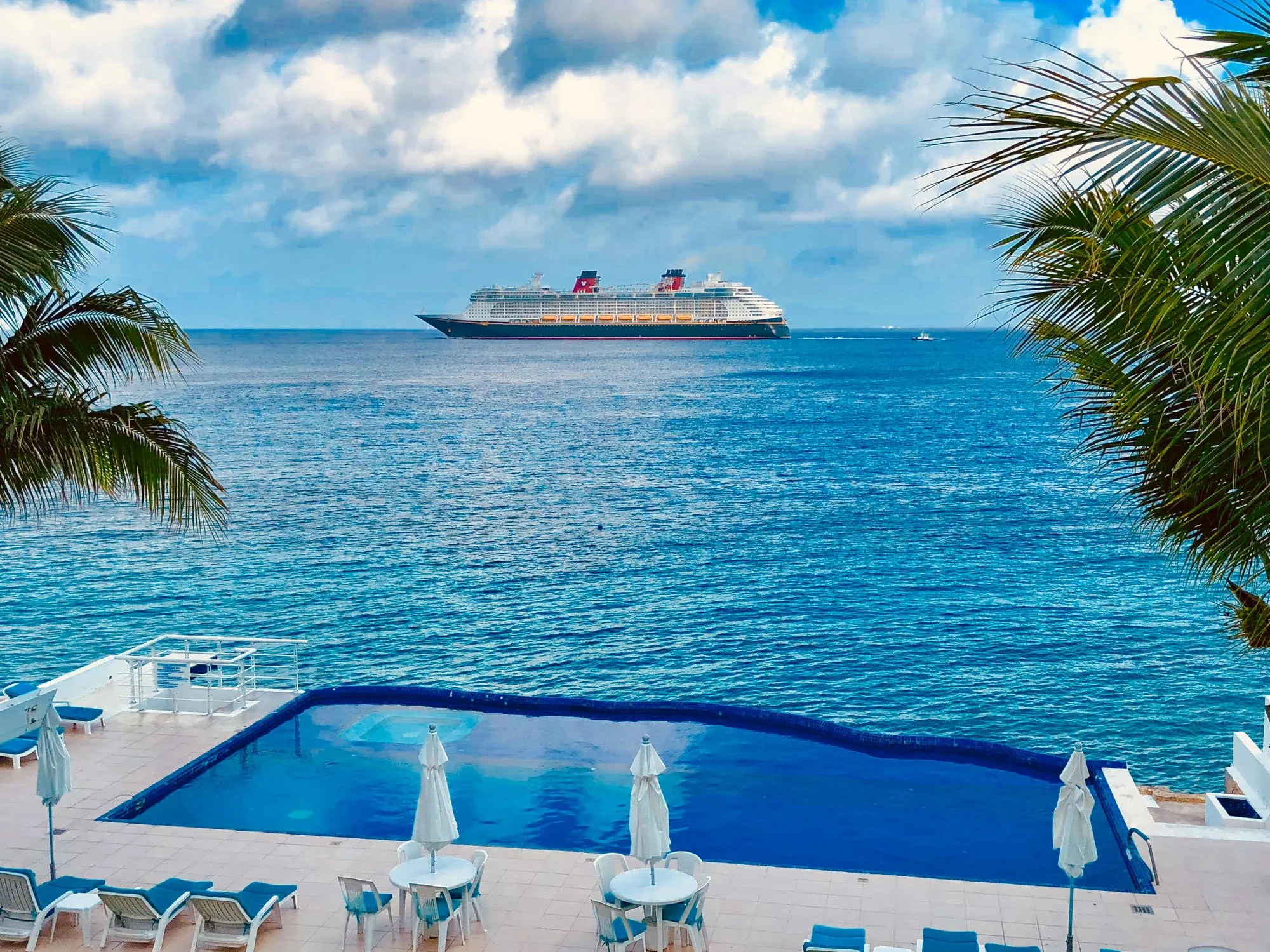 Swimming pool area with lounge chairs, umbrellas, and tables in the foreground, overlooking the ocean. A large cruise ship is on the water in the distance, with palm trees on either side of the image.