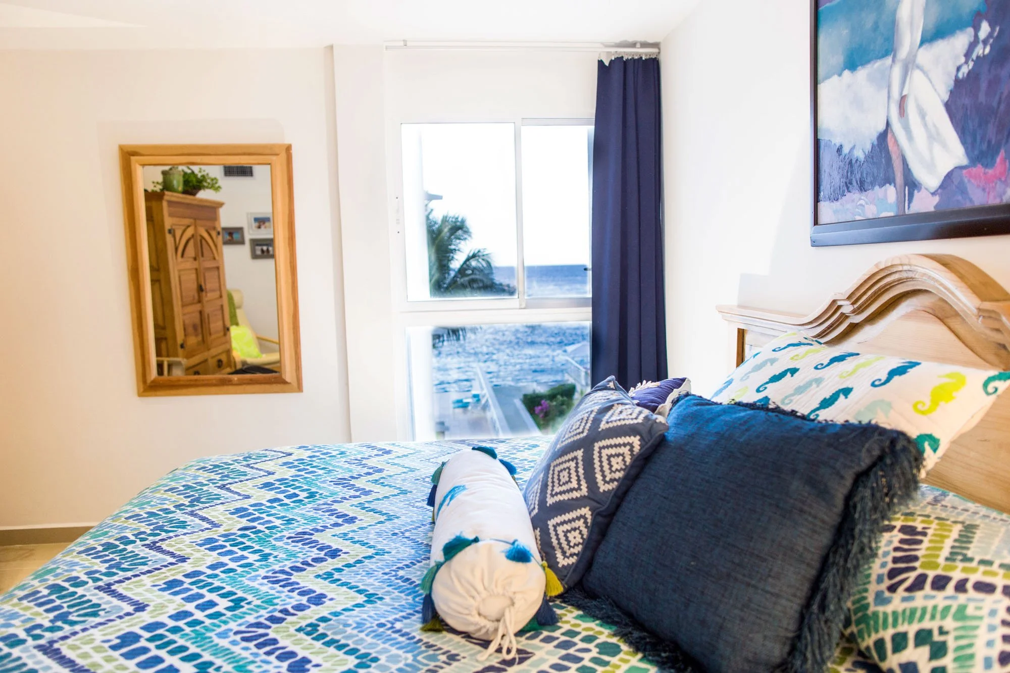A bedroom with a bed covered in a colorful, patterned quilt and decorative pillows, a window with a view of the ocean and palm trees, a wooden headboard, a wall-mounted painting, and a mirror reflecting part of the room.