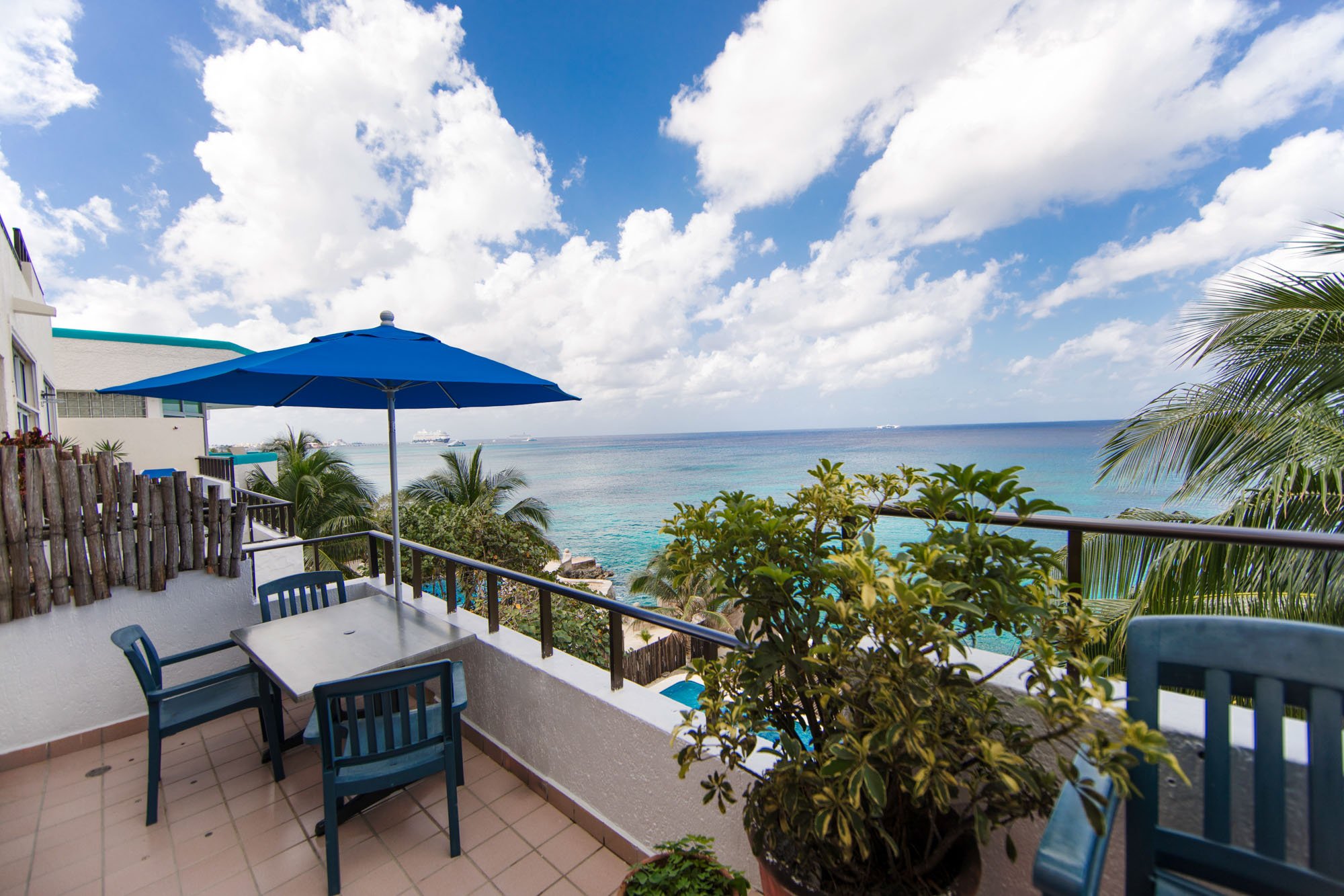 Balcony with blue chairs, table, umbrella, and plants overlooking ocean with cruise ships and a partly cloudy sky.