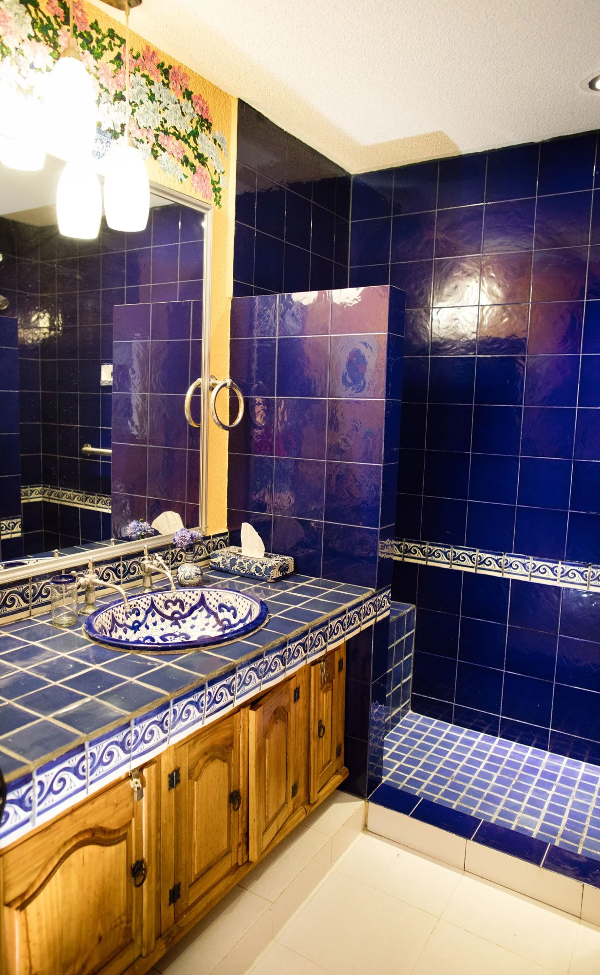 A bathroom with blue tiled walls and a shower area. Wooden vanity with a blue patterned sink, tissue box, and decorative items. Mirror with hanging lights above the sink. Floral border above the mirror.